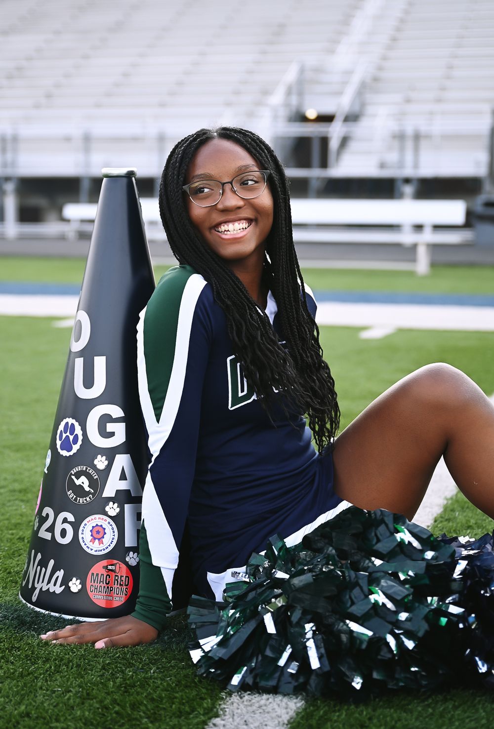 Cheerleader in uniform sitting on football field grass, smiling, holding a decorated megaphone with pom-poms.