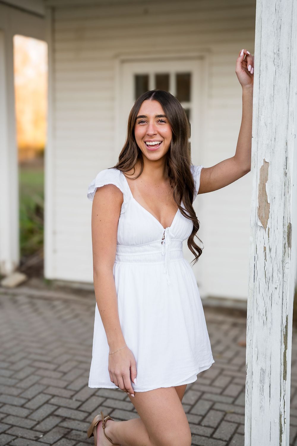 Young woman in white mini dress smiling and posing against a rustic white column outdoors.