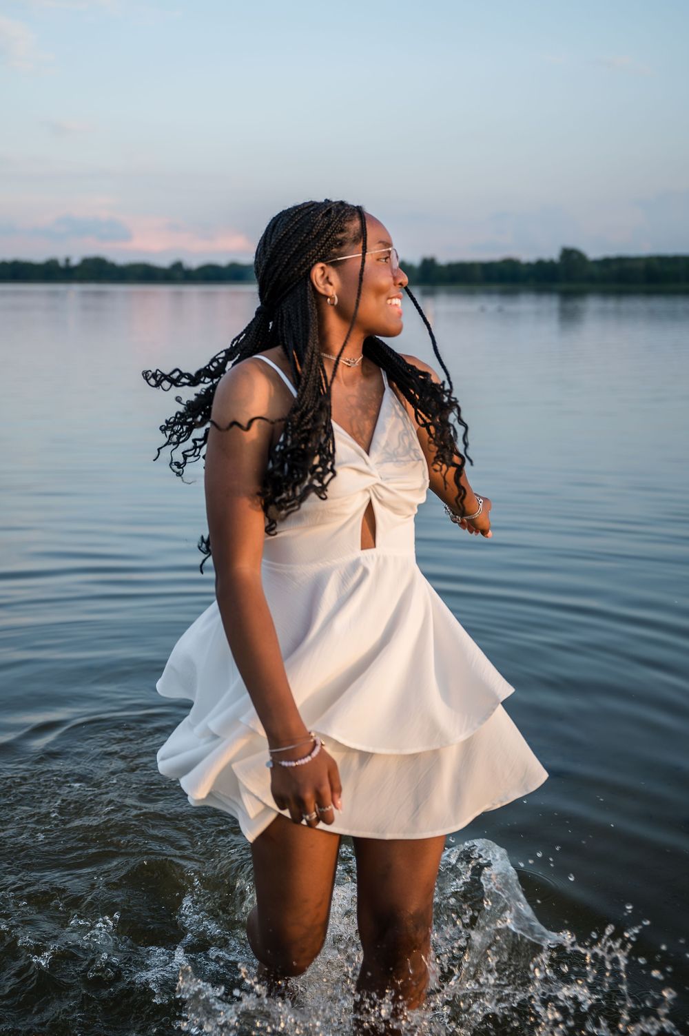 Young Black woman in white dress standing in lake water at sunset, braided hair flowing in the wind.