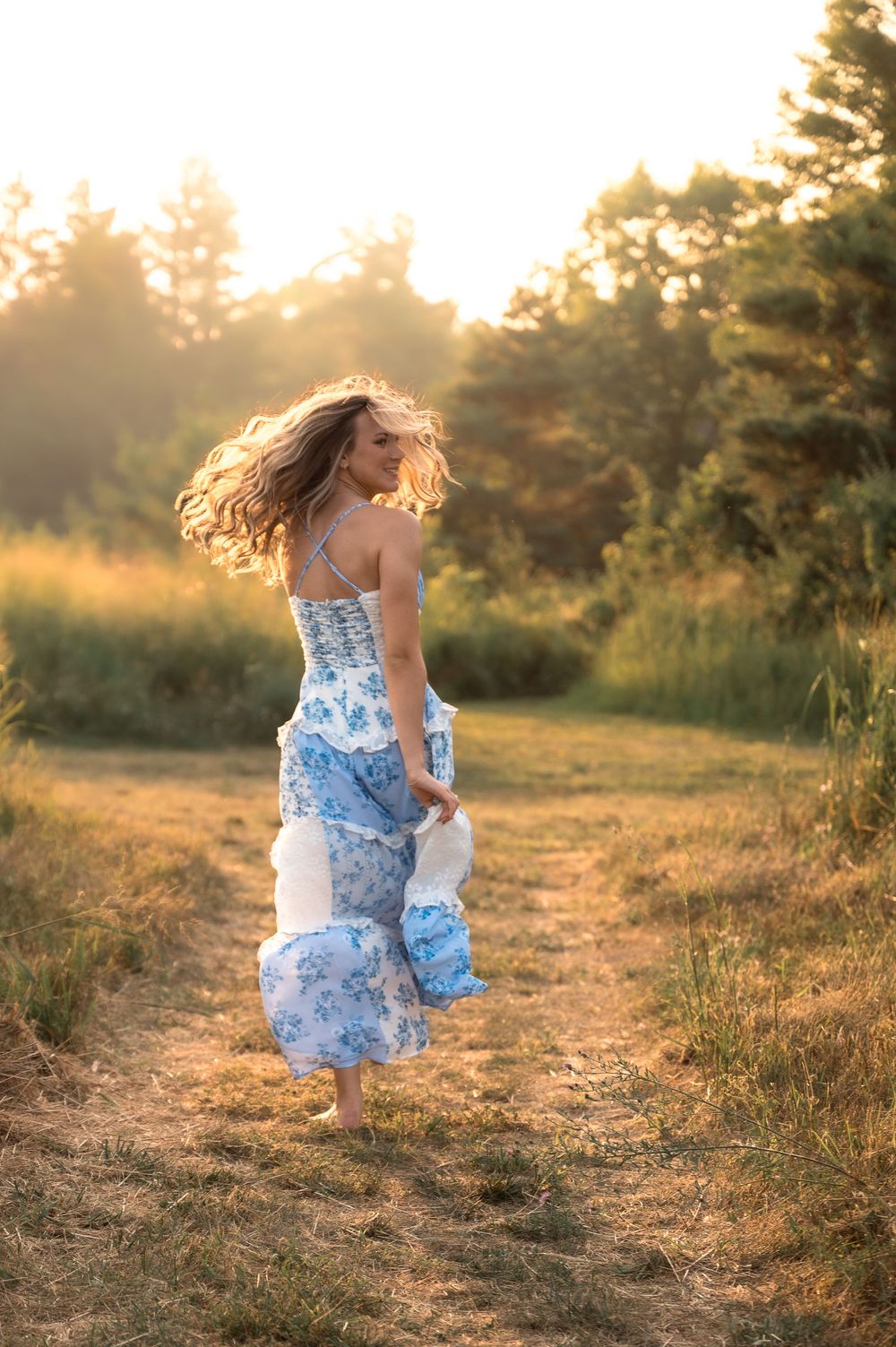 Woman in blue floral dress twirling on dirt path in sunlit meadow at golden hour.