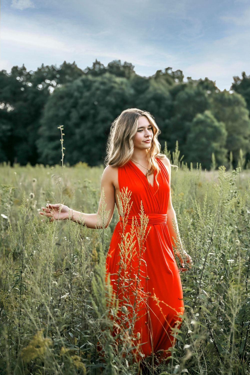 Young blonde woman in red dress walking through green field with wildflowers and trees in background.