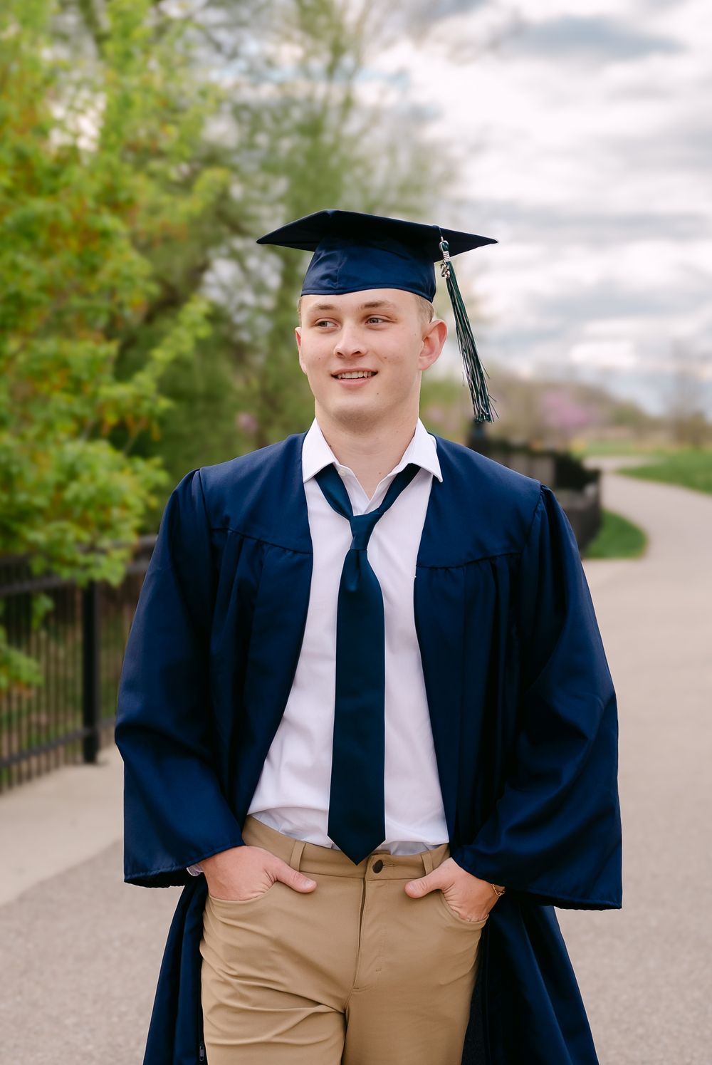 Young man smiling in navy graduation cap and gown with khaki pants, posing outdoors on a tree-lined path.