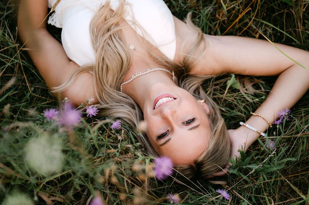 Smiling blonde woman lying in green grass surrounded by purple wildflowers in golden light.