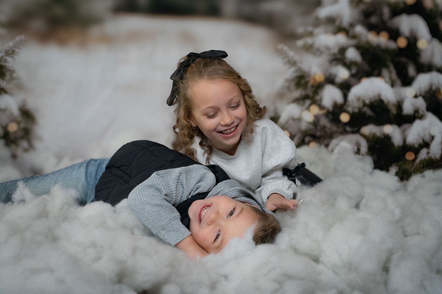 Two happy children playing in snow near a Christmas tree with bokeh lights, holiday winter scene.