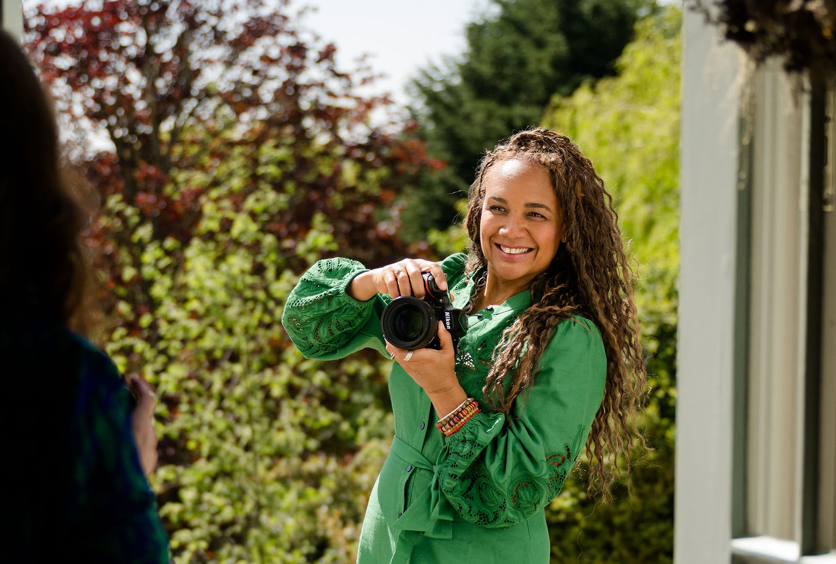 A smiling young woman in a green shirt holds a camera outdoors surrounded by lush green trees.
