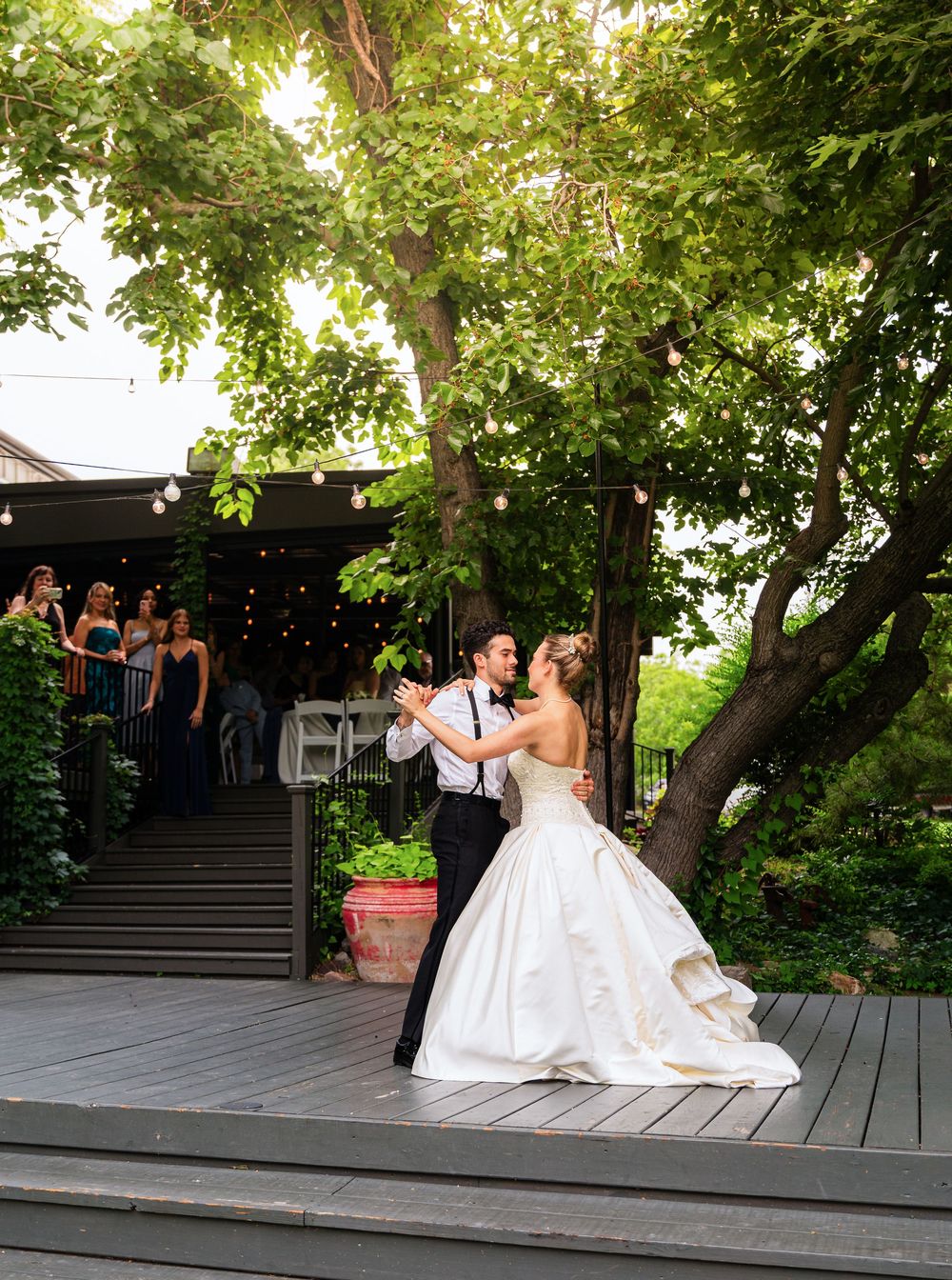 Bride and groom sharing romantic first dance on outdoor wooden deck surrounded by string lights and guests.