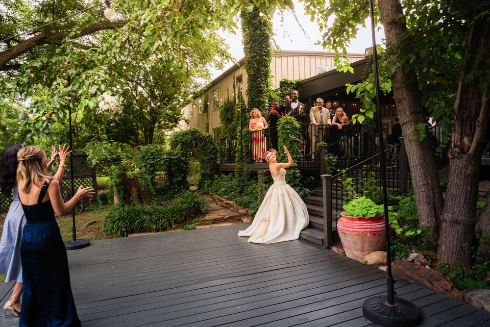 Bride in white gown tossing bouquet on outdoor deck surrounded by lush greenery at elegant wedding venue.