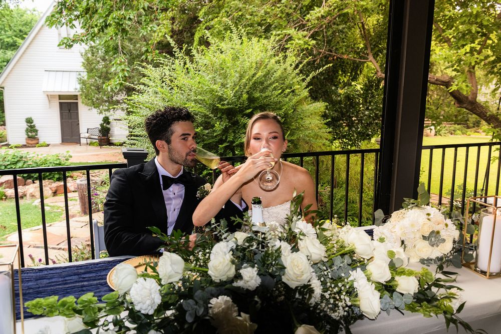 Bride and groom sharing a toast at sweetheart table adorned with white floral arrangements at outdoor wedding.