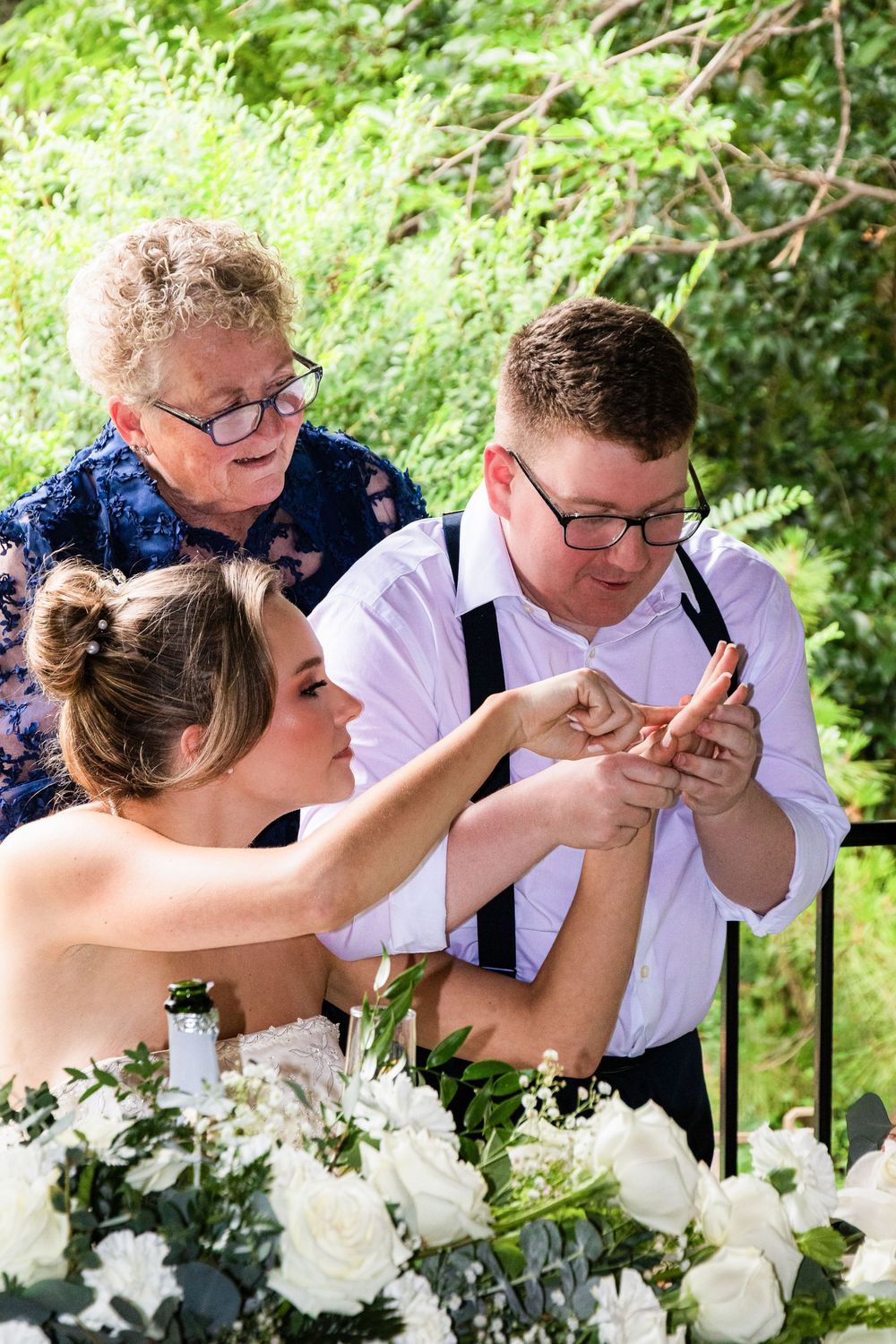 Bride placing wedding ring on groom's finger during outdoor ceremony with officiant, surrounded by white roses.
