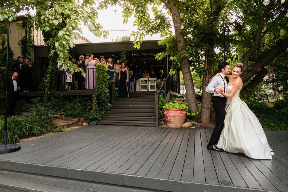 Bride and groom share a romantic dip kiss on outdoor deck while wedding guests watch from steps surrounded by lush greenery.