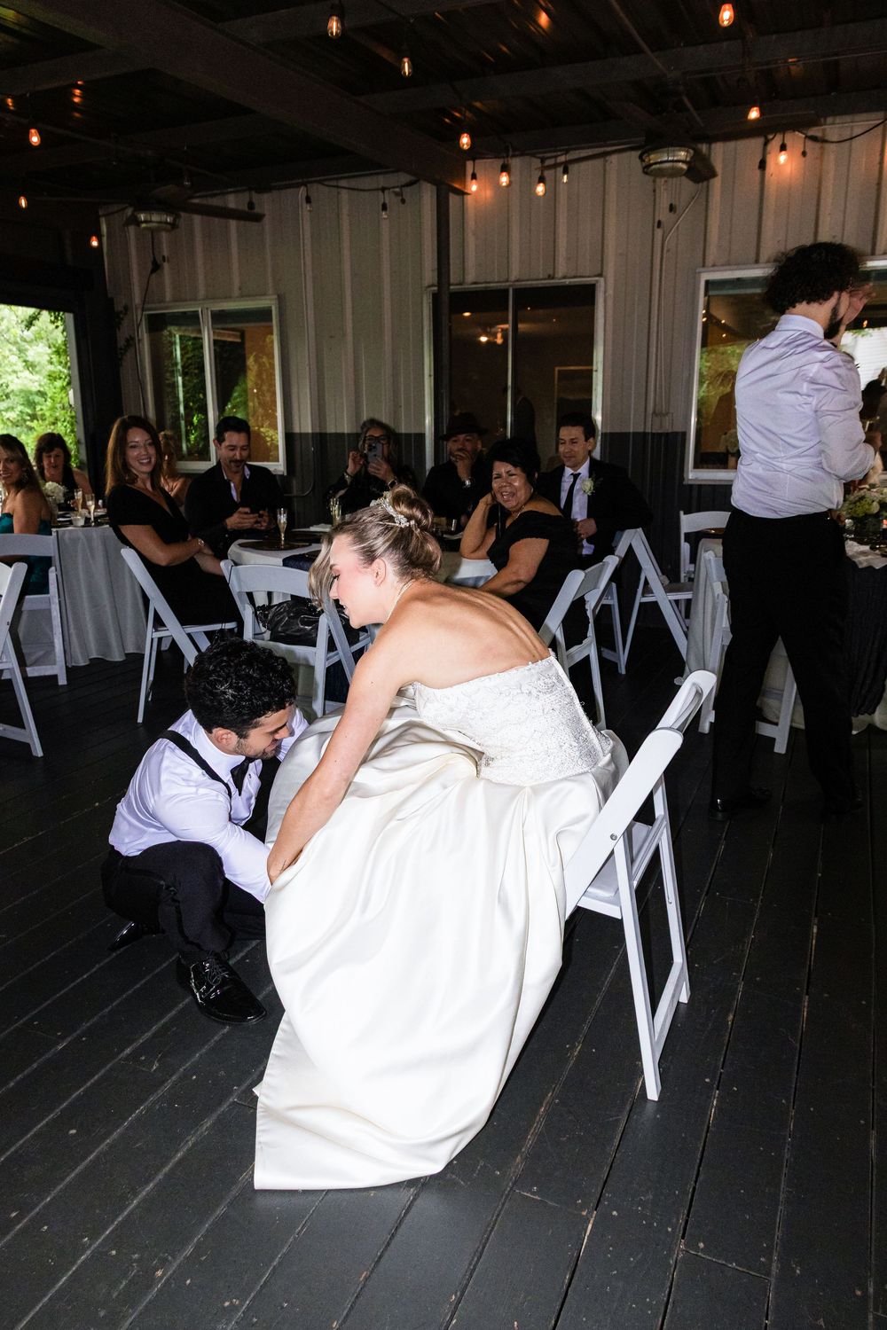 Bride in white gown seated as groom removes garter at indoor wedding reception with guests watching in background.