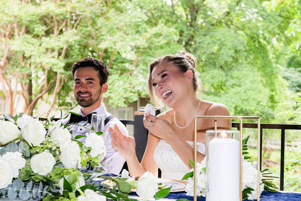 Bride and groom smiling and laughing at sweetheart table adorned with white floral arrangements at wedding reception.