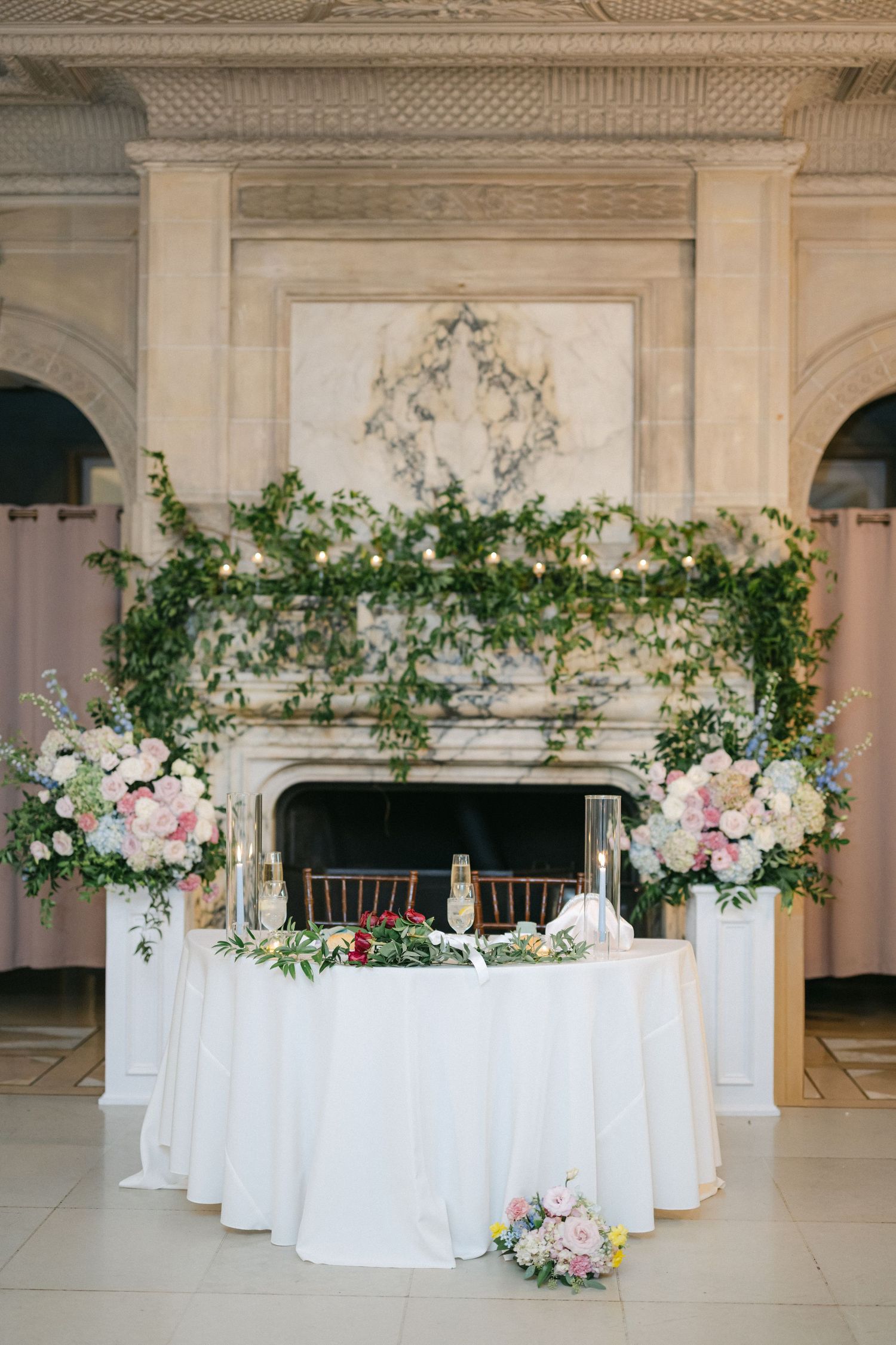 Elegant wedding sweetheart table with white linen, pink floral arrangements, and lush greenery draped over a grand fireplace.