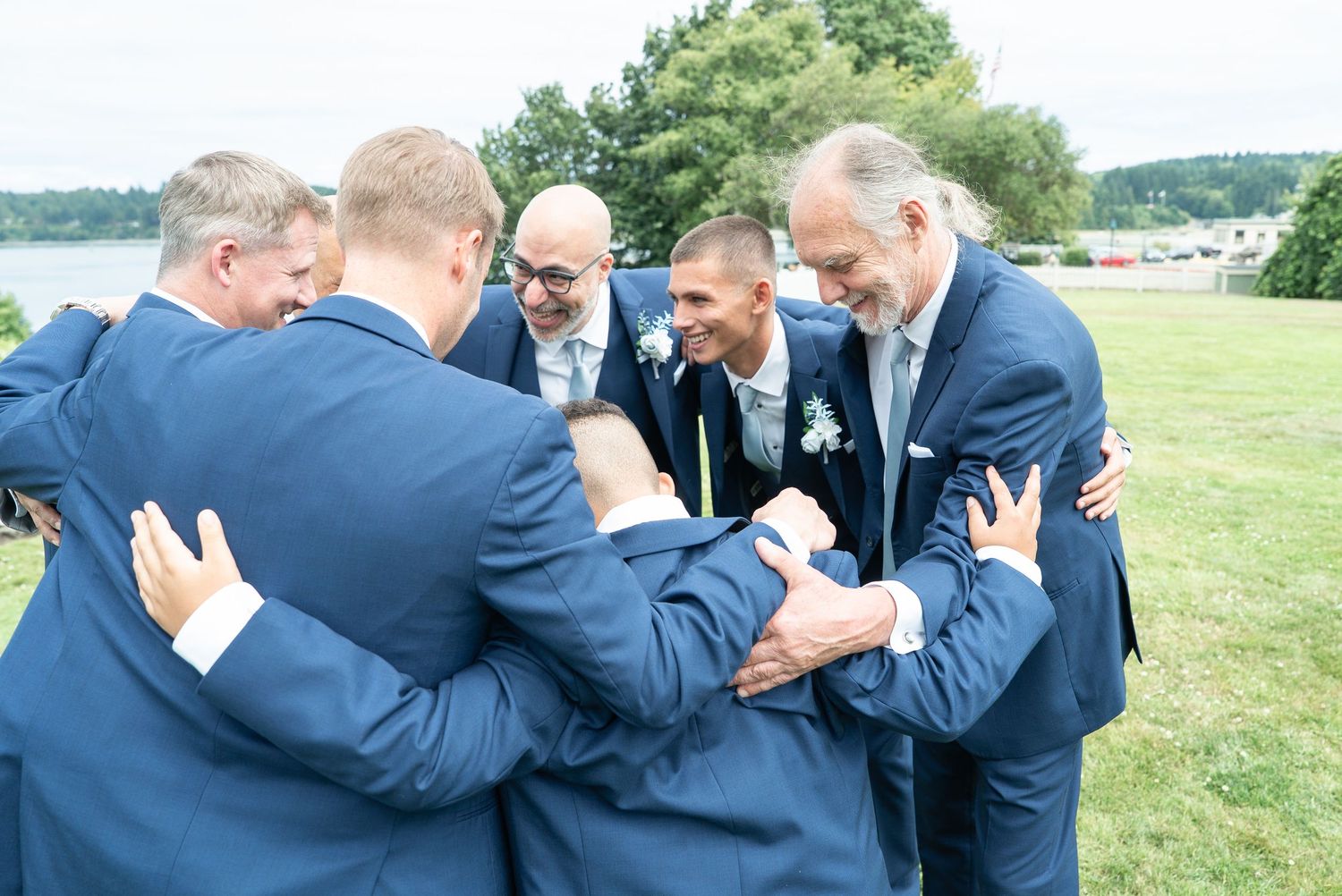 Group of men in navy suits sharing a warm embrace outdoors at a wedding celebration near a waterfront.