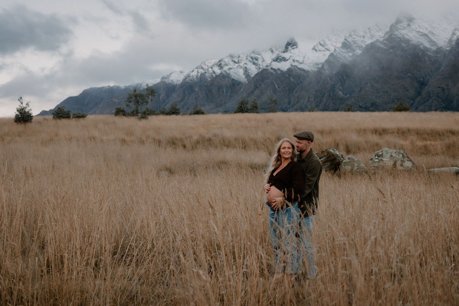 Couple embracing in golden field with snow-capped mountains backdrop during moody overcast day.
