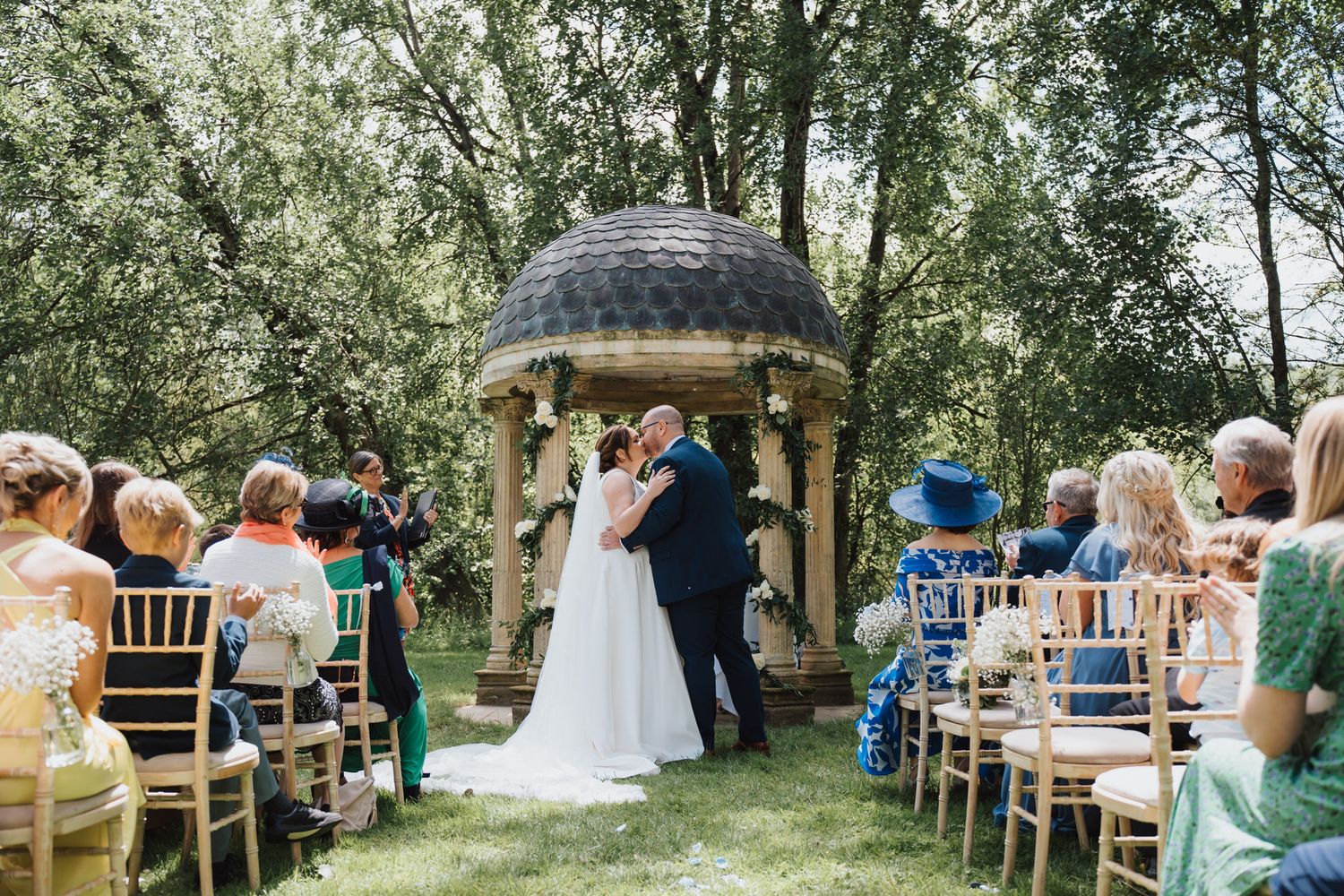 Couple shares first kiss at outdoor wedding ceremony beside stone gazebo, guests seated on wooden chairs in lush garden.