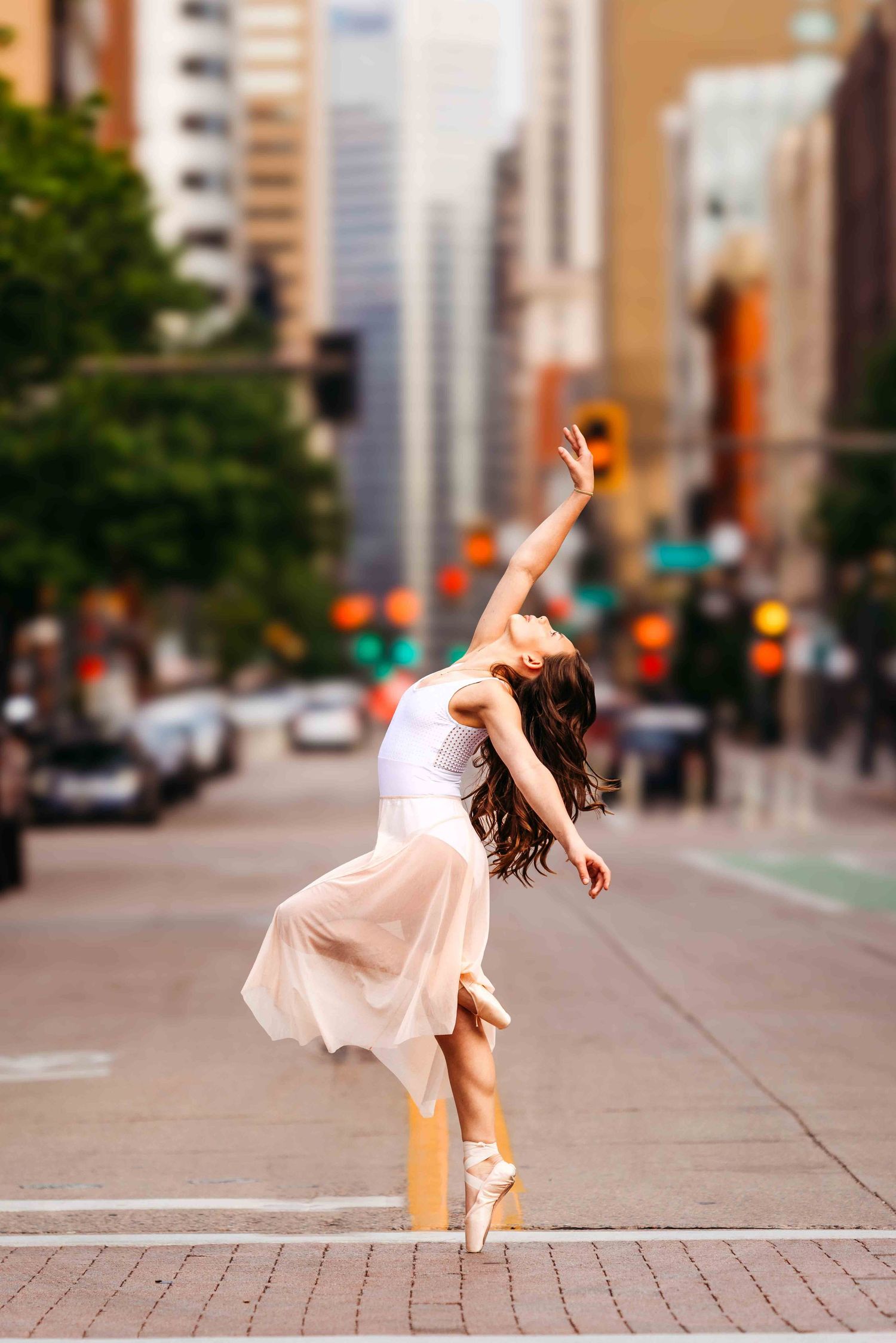 A graceful ballerina in a white top and blush skirt poses en pointe on a city street with blurred urban backdrop.