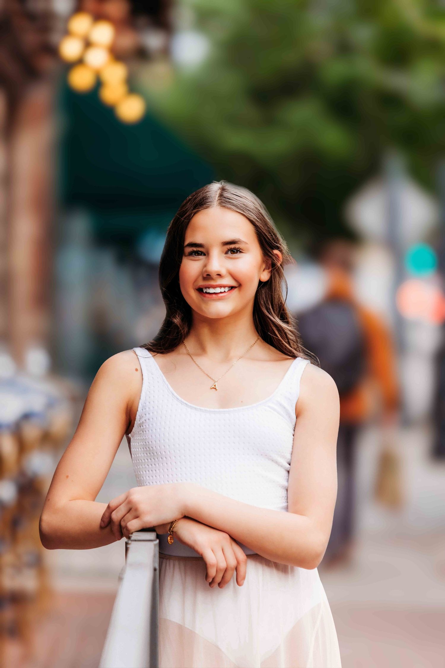 Smiling young woman in white dress leaning on railing on a blurred urban street background.
