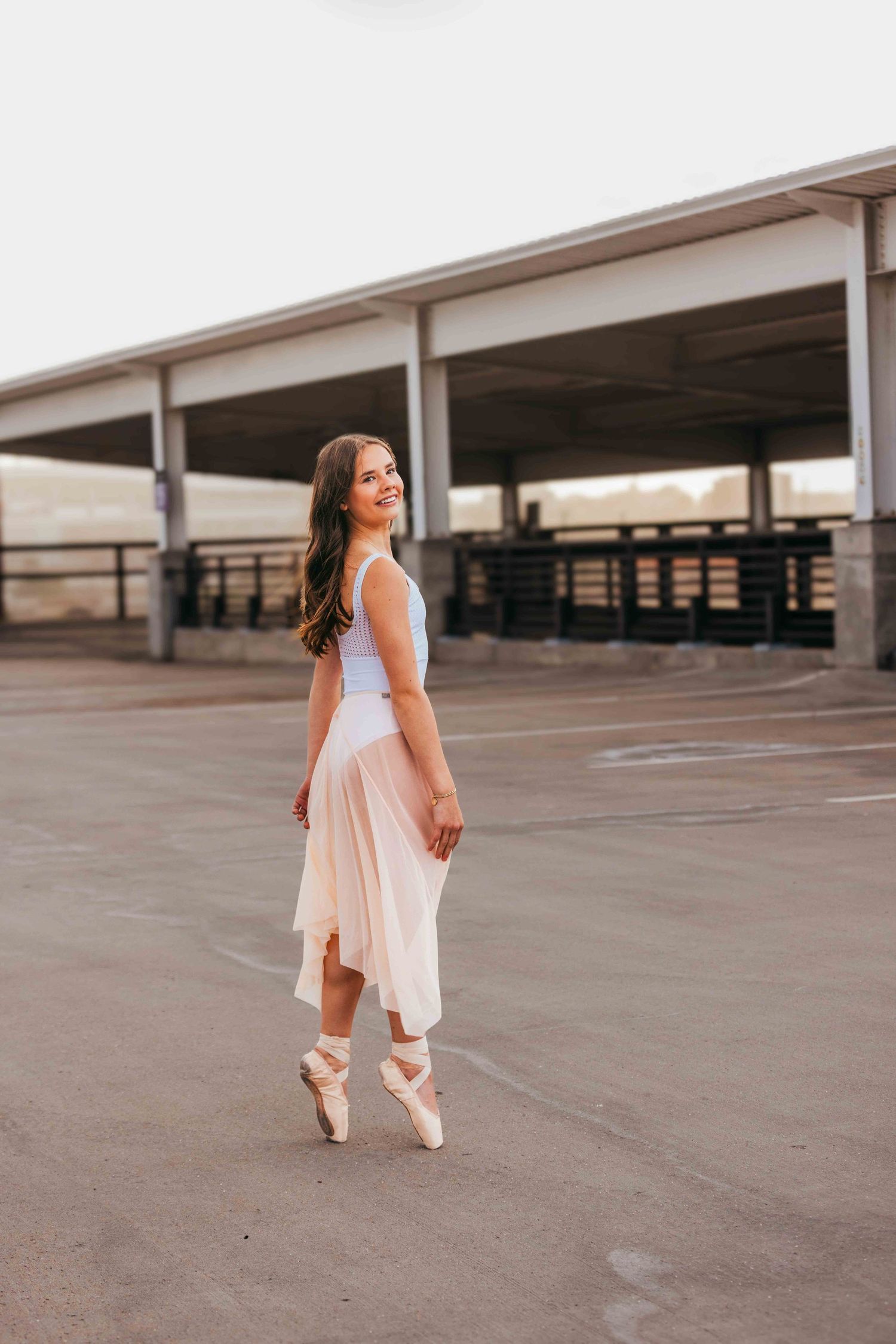 A ballerina in a pink tulle skirt and white leotard poses on pointe in an urban parking structure.