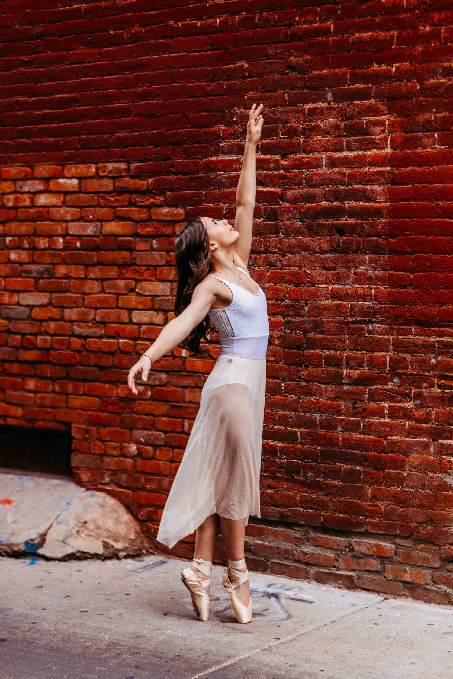 Graceful ballerina in white leotard and sheer skirt poses en pointe against a red brick wall outdoors.