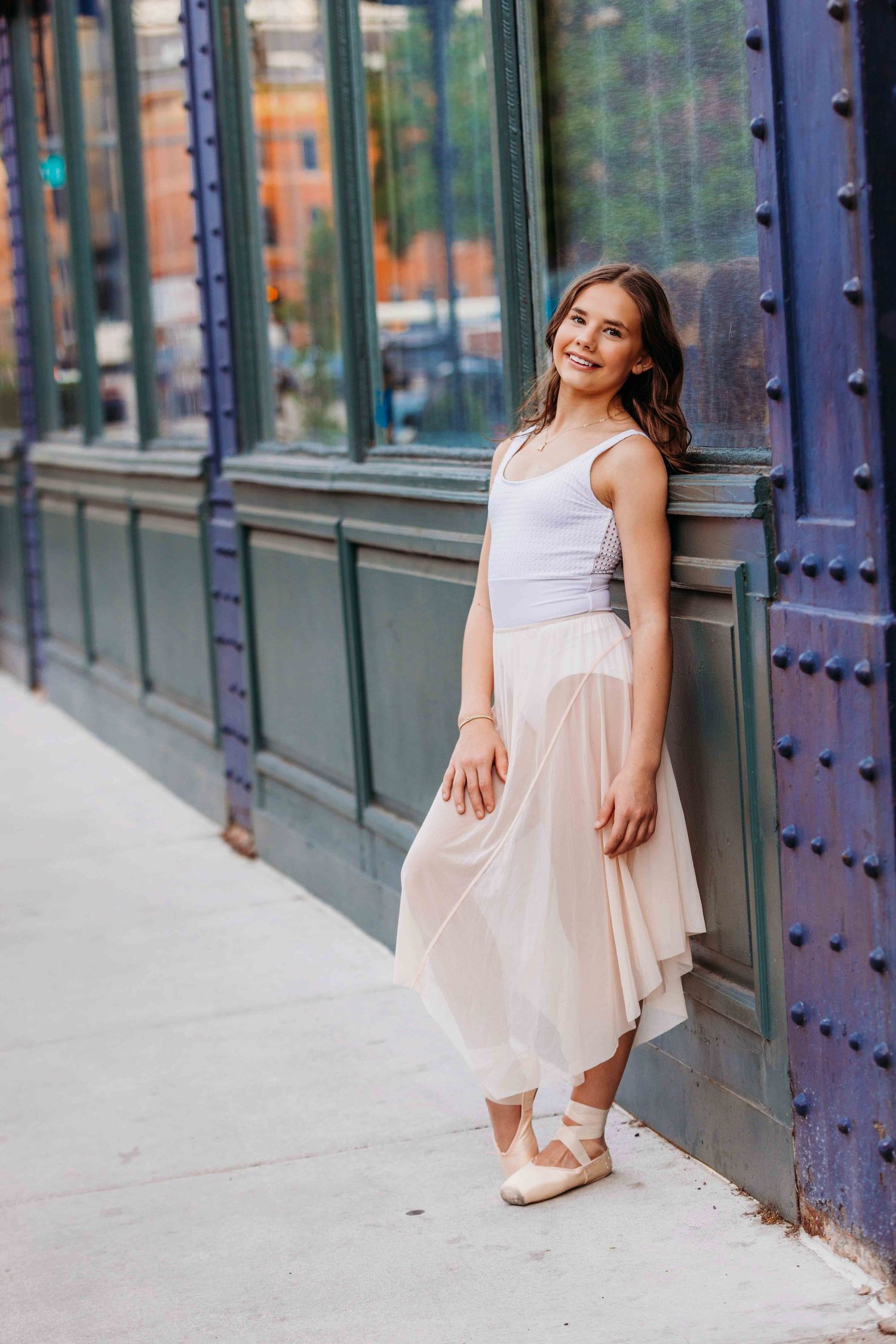 Young ballerina in white leotard and sheer skirt leaning against a decorative building exterior.