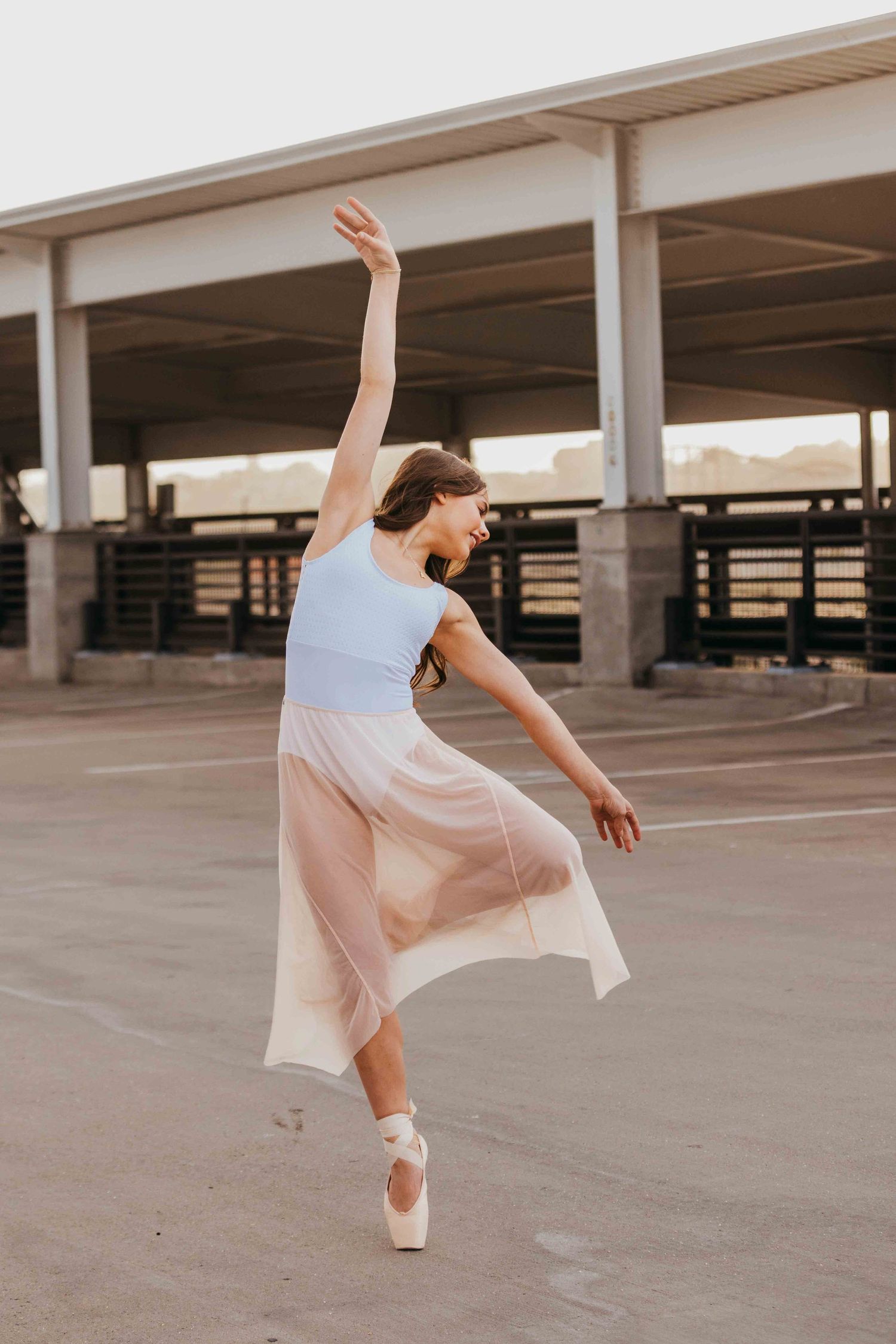 A graceful ballerina in a white leotard and pink tulle skirt performs en pointe outdoors near a parking structure.