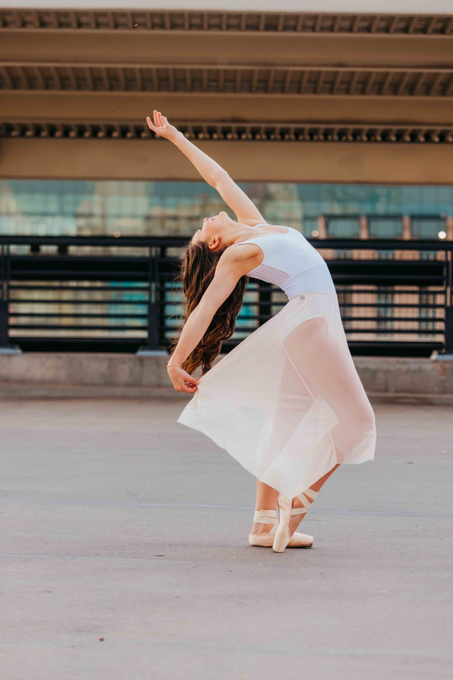 Graceful ballerina in white tutu and pointe shoes performs elegant arabesque outdoors in urban setting.