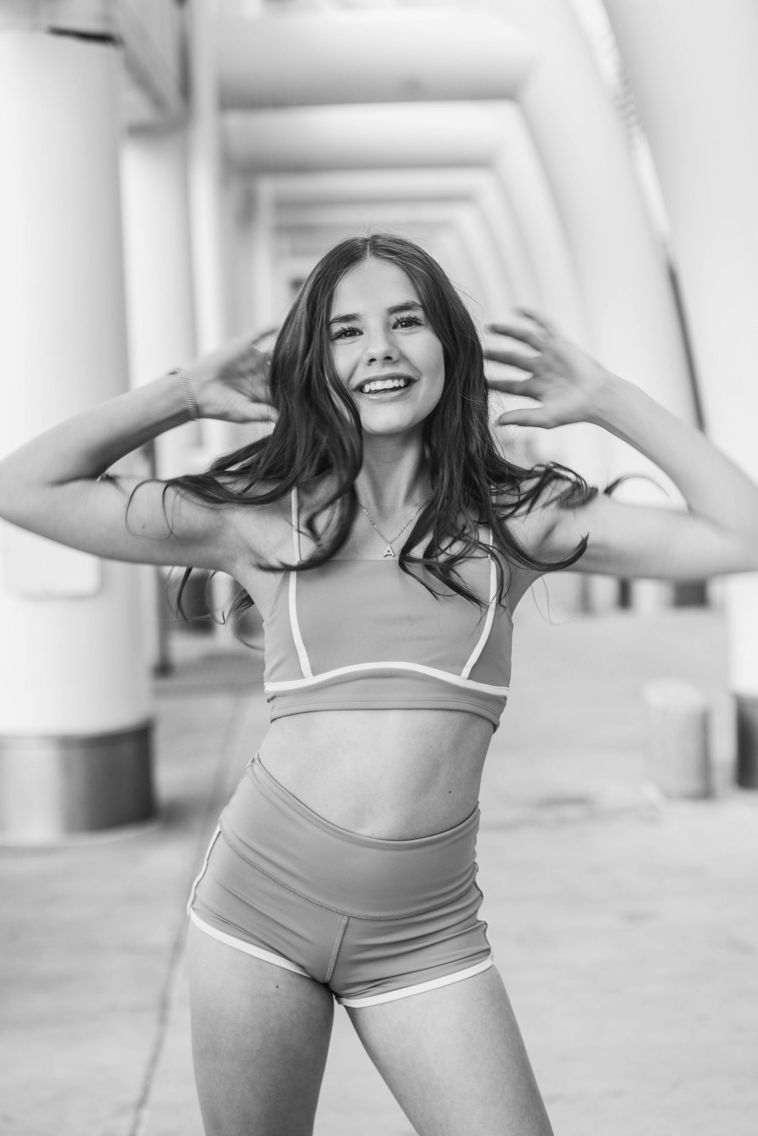 Young woman in athletic crop top and shorts smiling outdoors near columns in black and white photo.