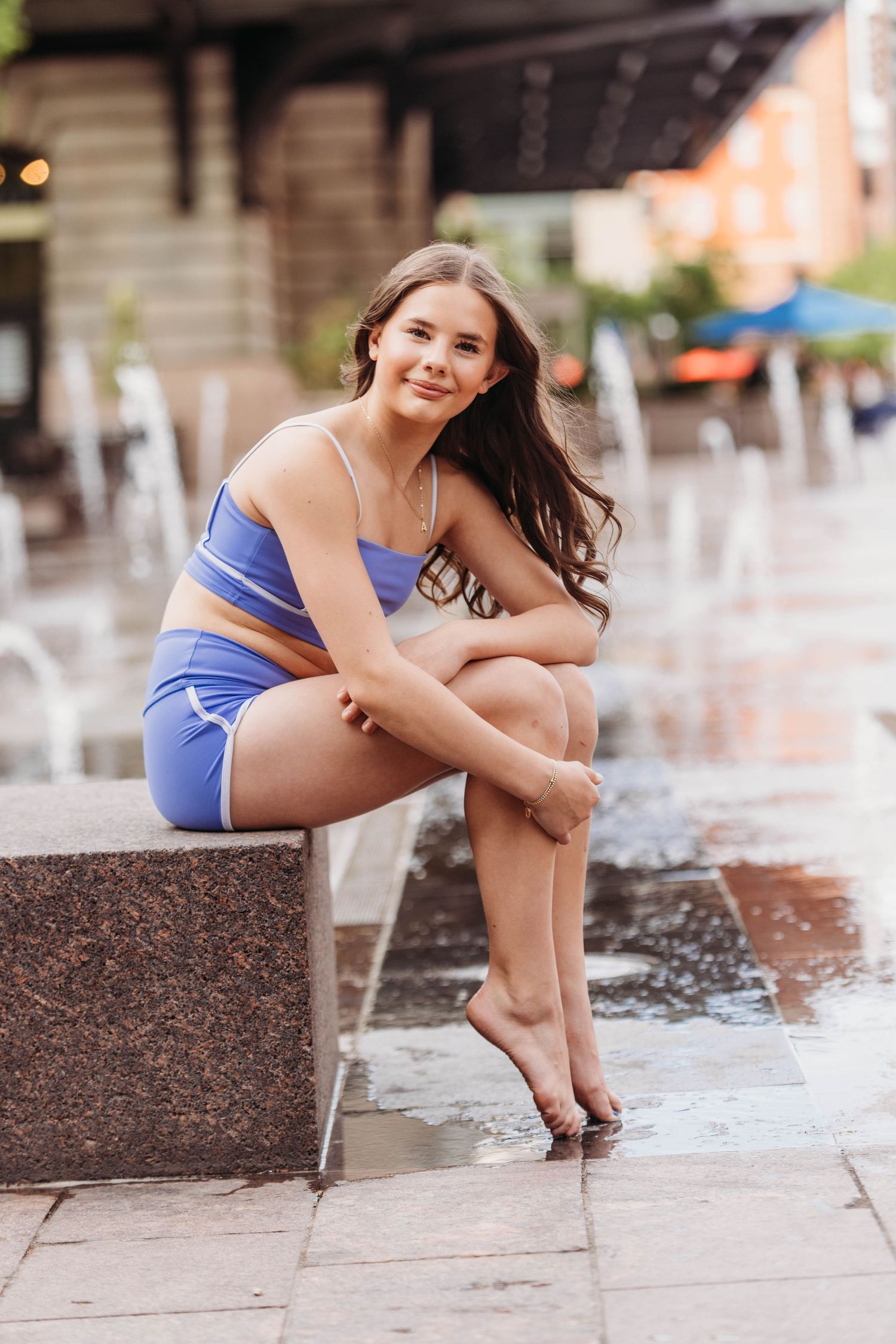 Young woman in blue athletic set sitting by urban fountain, smiling at camera on sunny day.