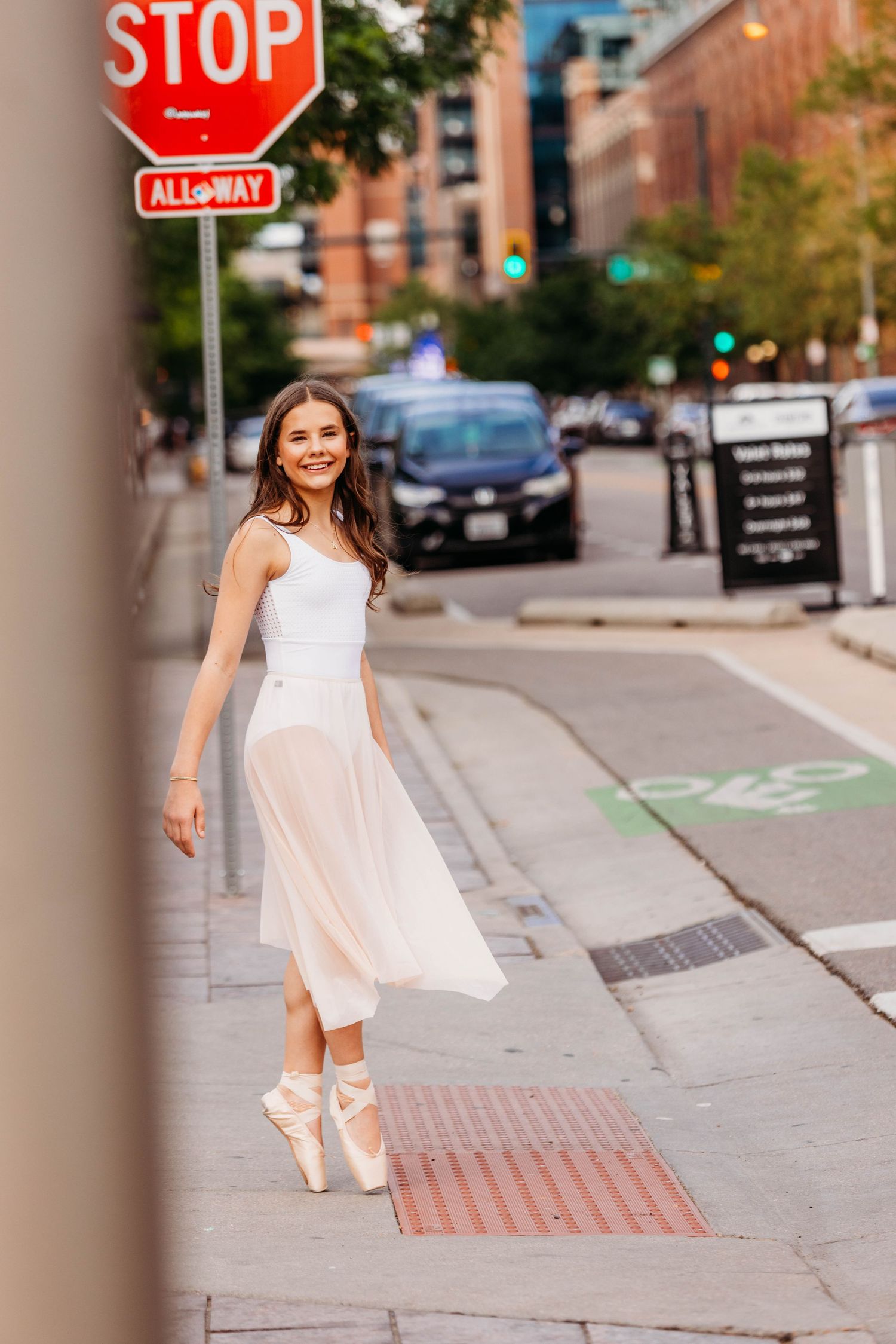 A smiling ballerina in white dress and pointe shoes poses gracefully on a city sidewalk near a stop sign.