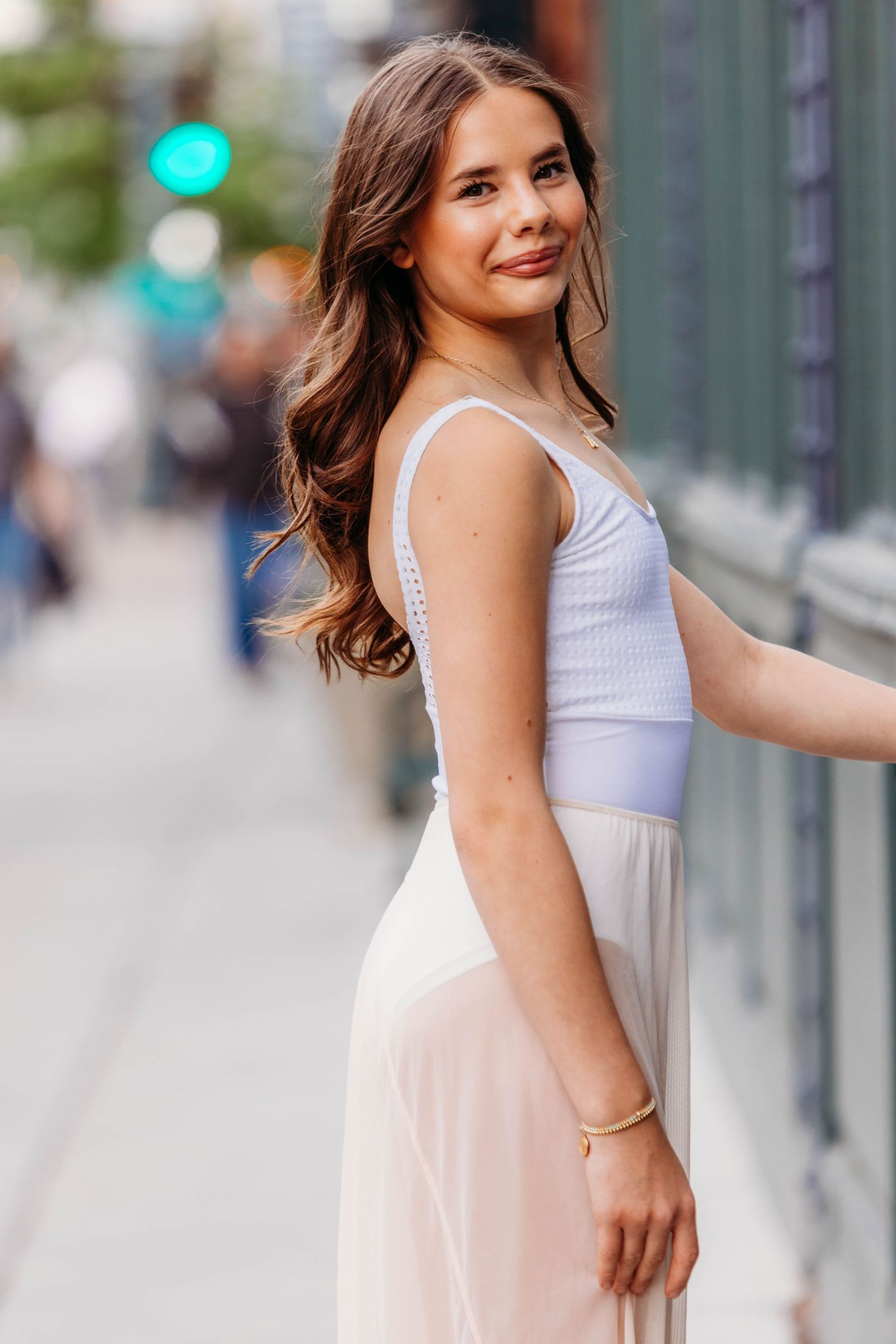 Young woman in white top and blush maxi skirt posing on urban sidewalk, smiling over shoulder.