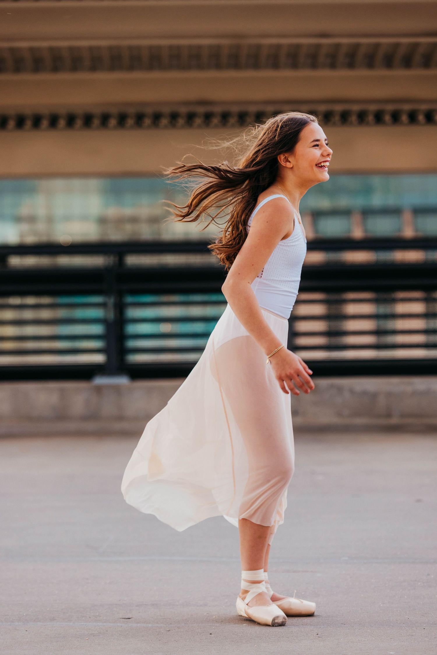 A smiling ballerina in white top, pink skirt, and pointe shoes dancing outdoors with hair flowing in the wind.