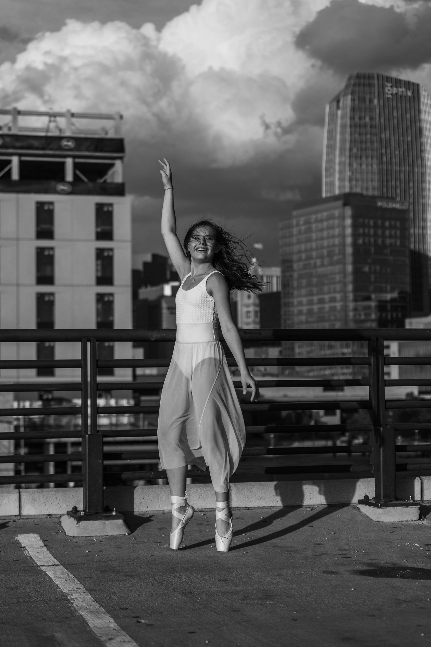 Black and white photo of a ballerina en pointe on a rooftop with city skyline and dramatic clouds behind her.