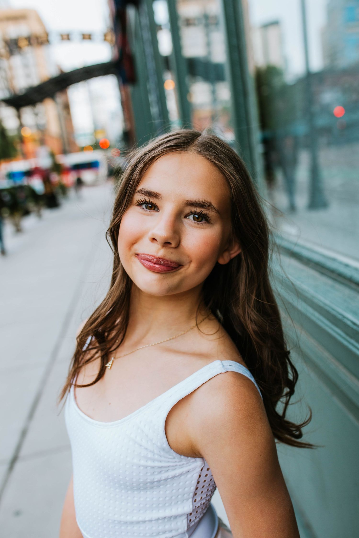 Young woman with long brown hair smiling in white top, posing on urban city sidewalk near glass storefront.