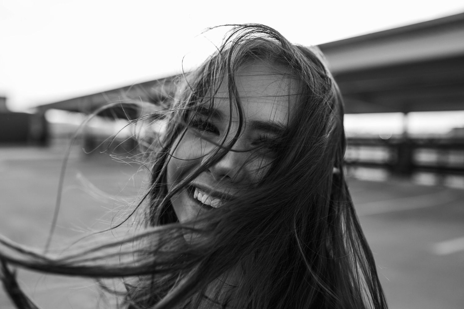 Black and white photo of a smiling woman with wind-blown hair outdoors near a highway overpass.