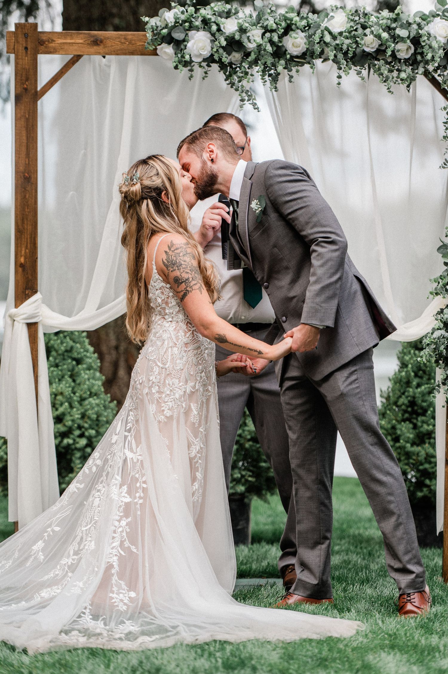 Bride and groom share first kiss under rustic wooden arch with white draping and greenery during outdoor wedding ceremony.