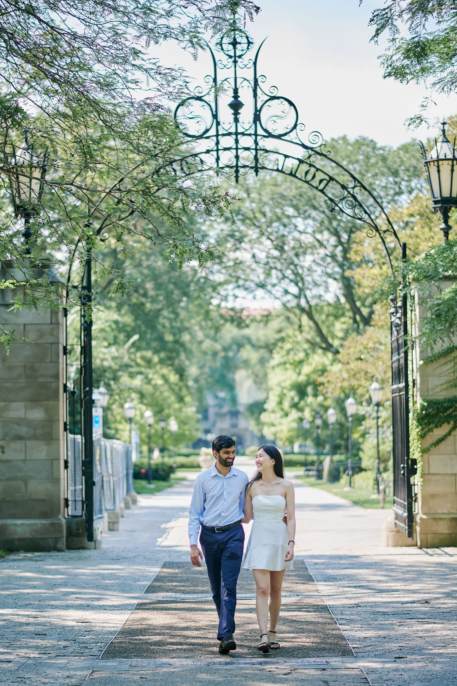 Couple walking hand-in-hand through an ornate iron gate archway surrounded by lush green trees on a sunny day.