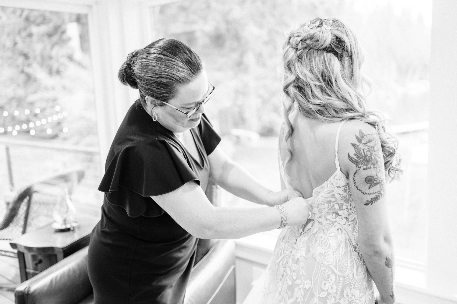 Black and white photo of a woman helping a bride button her lace wedding dress near a bright window.