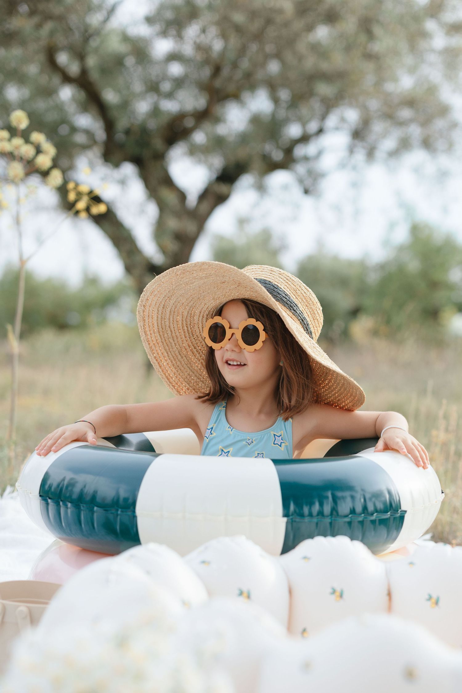 Young girl in straw hat and yellow sunglasses sitting in inflatable pool ring outdoors near a tree.