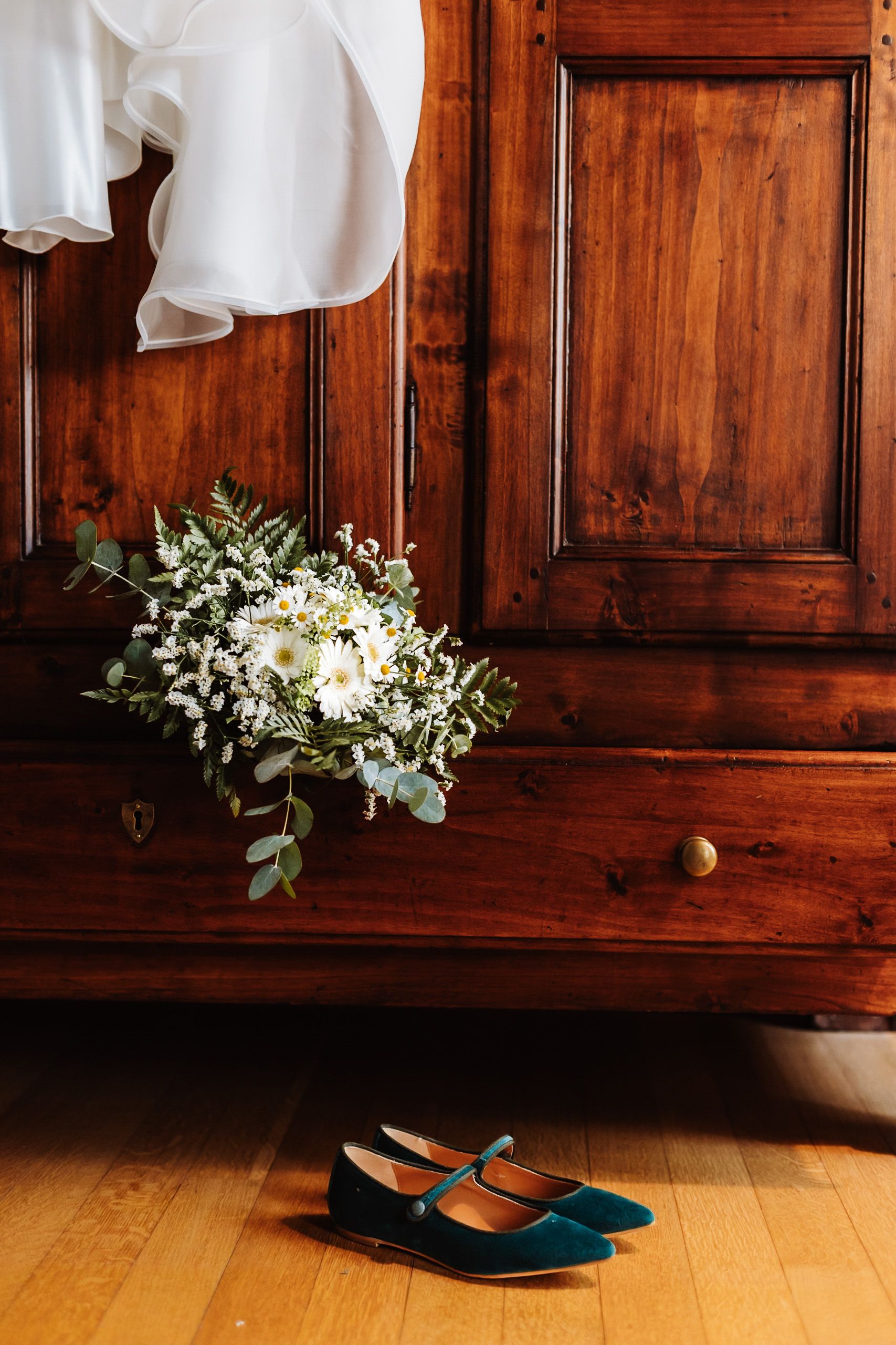 Bridal bouquet with white daisies and eucalyptus beside teal velvet shoes on wood floor near antique dresser.