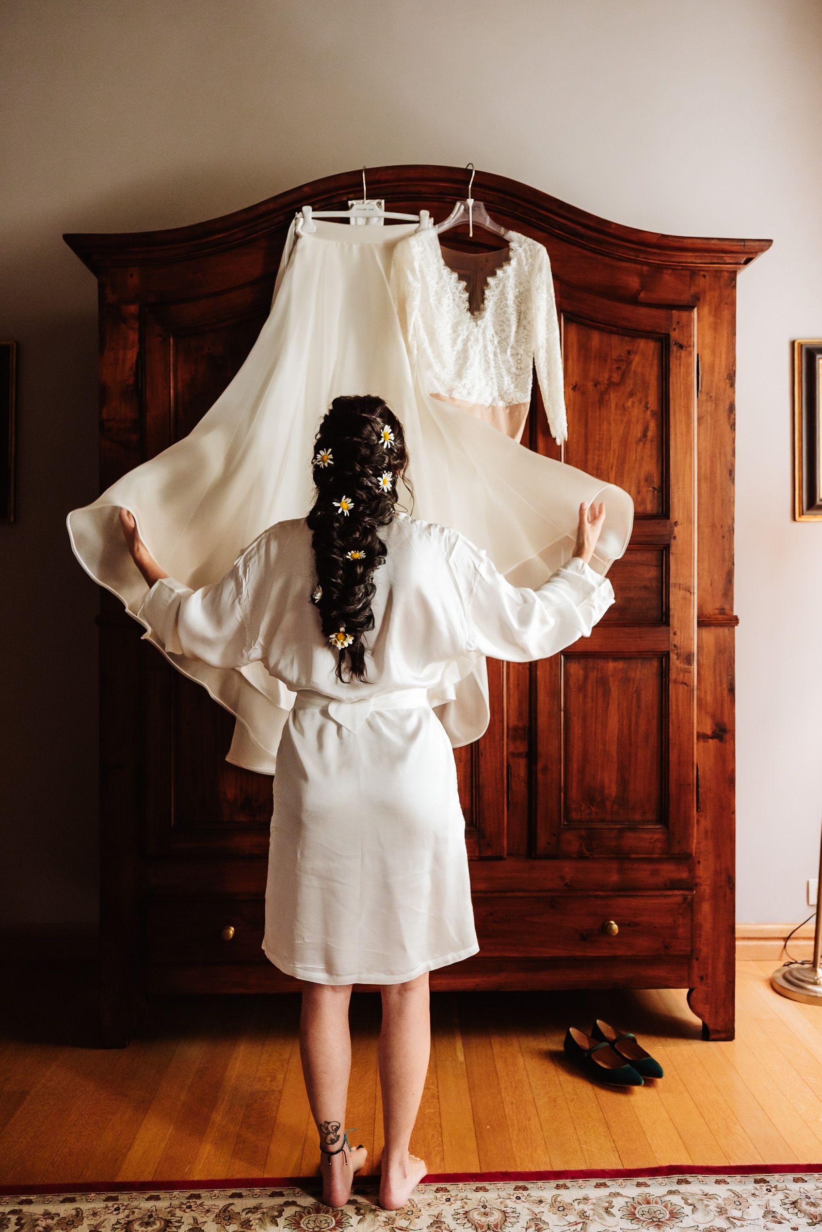 Bride in white robe admiring her wedding dress hanging on a wooden armoire before the ceremony.