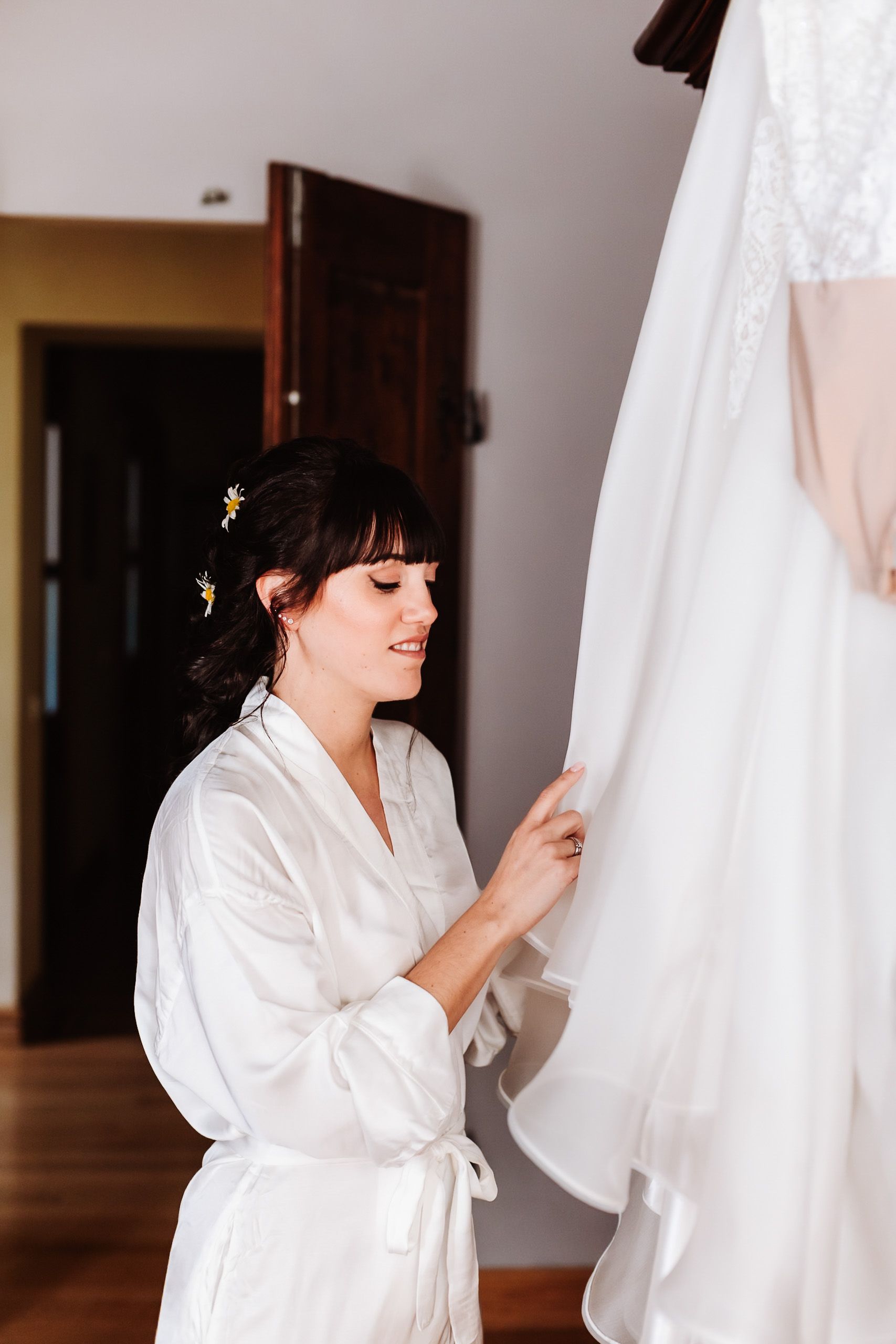 Bride in white satin robe admiring her wedding dress hanging in a bright room before the ceremony.