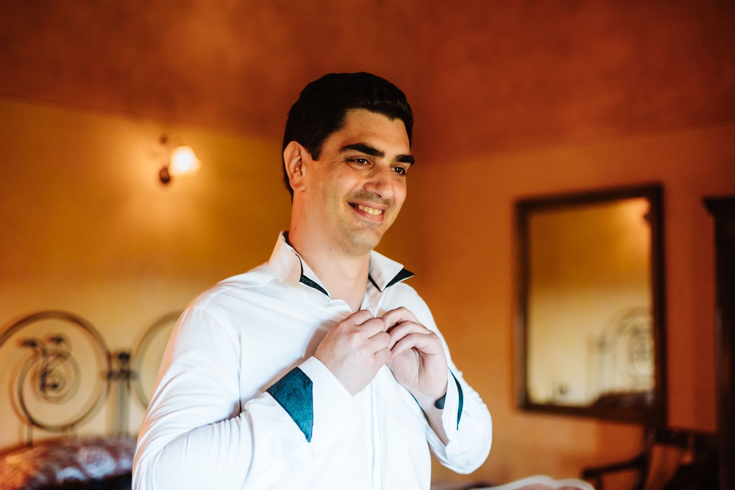 Groom in white shirt adjusting his teal tie while smiling in a warmly lit room before his wedding.