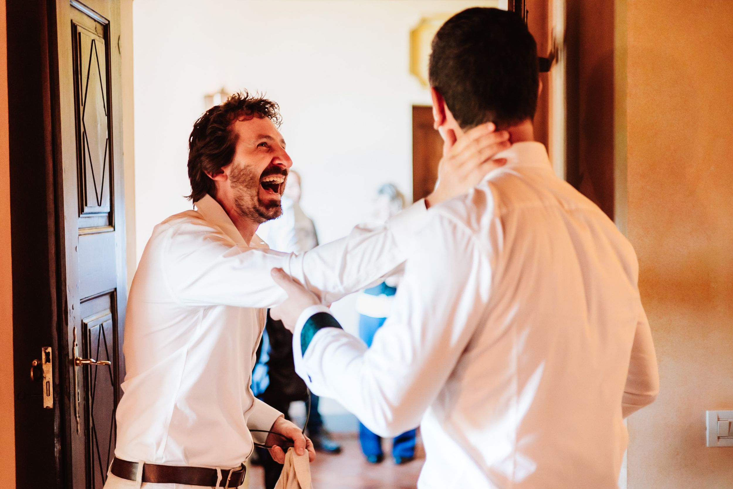 Two men in white shirts laughing and helping each other get ready for a wedding celebration.