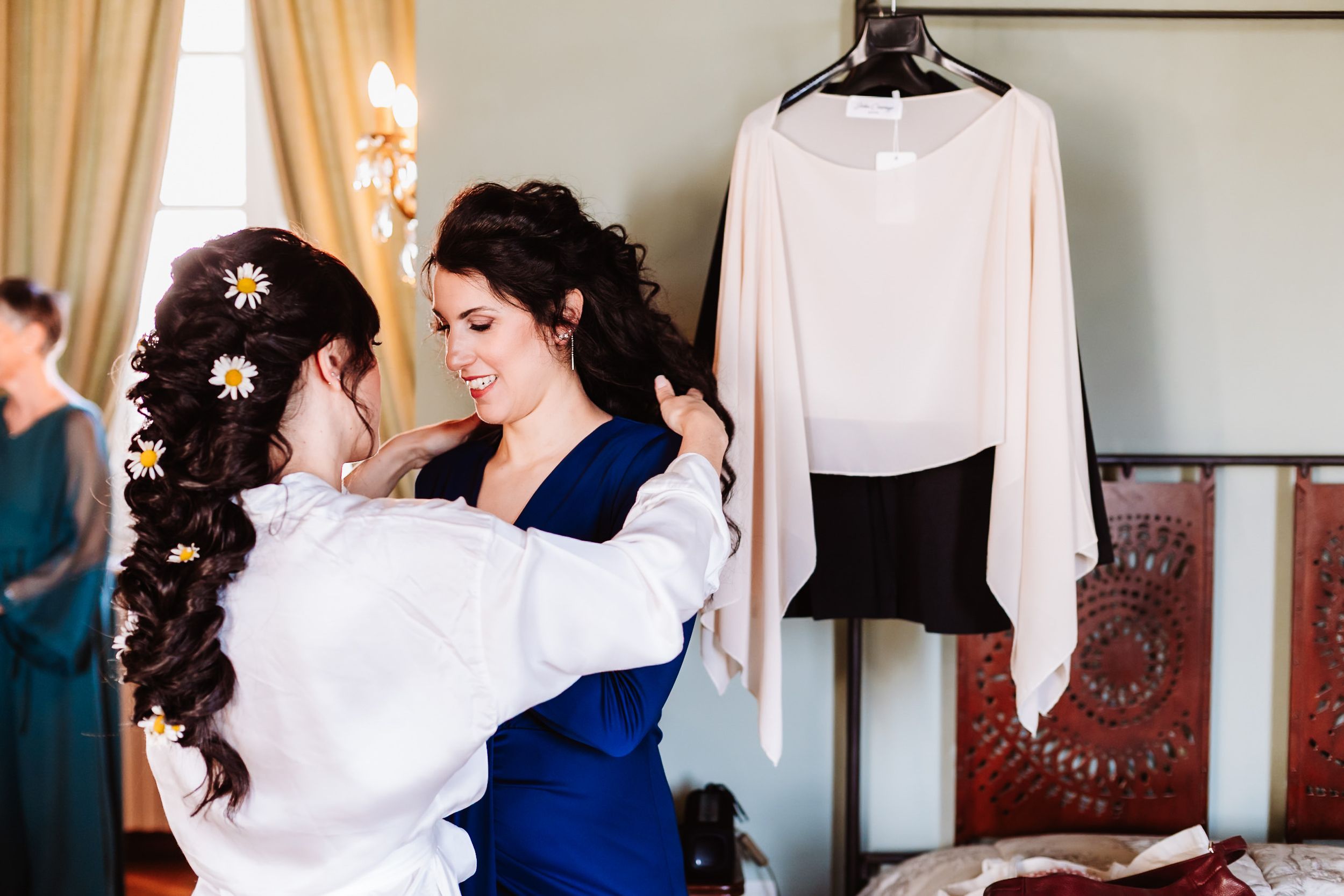Bridesmaid helping bride get ready, adjusting dress straps in elegant room with gown hanging in background.