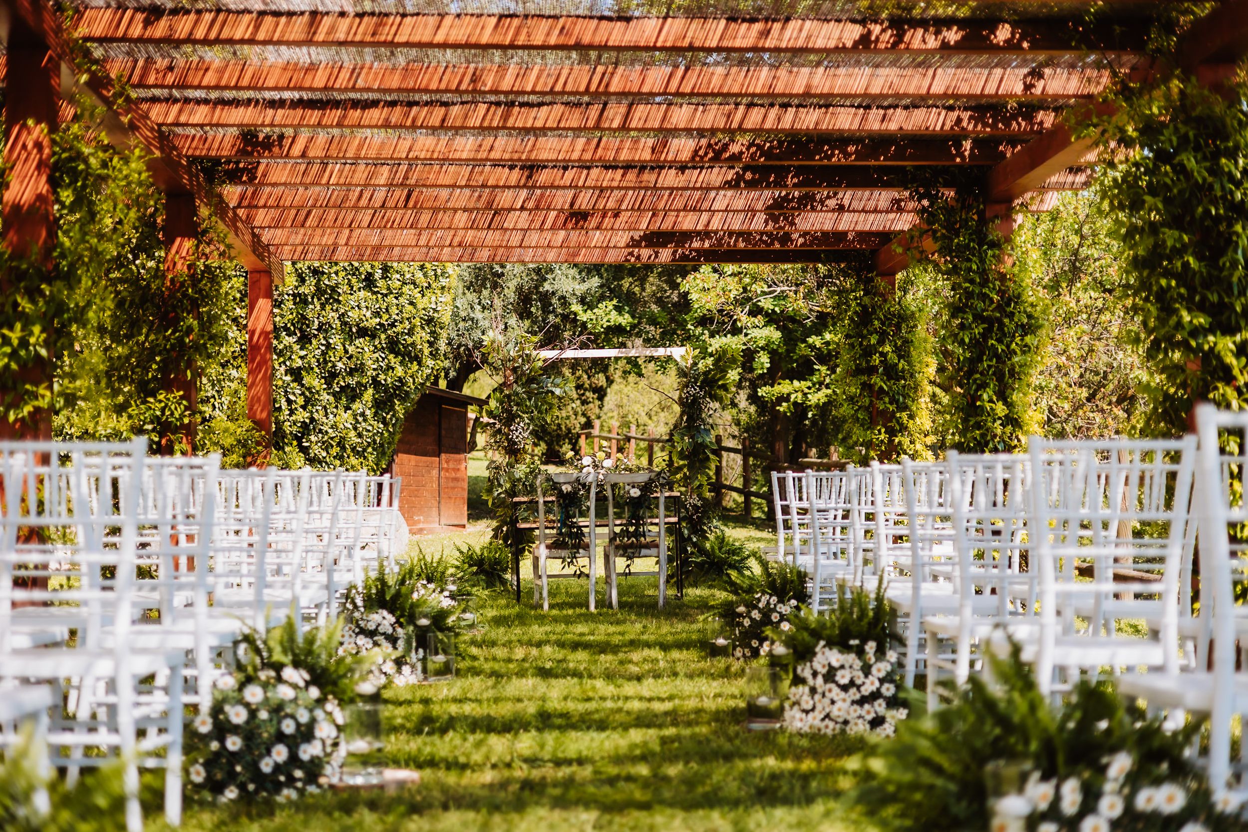 Elegant outdoor wedding ceremony setup with white chairs, floral aisle, and wooden pergola surrounded by lush greenery.