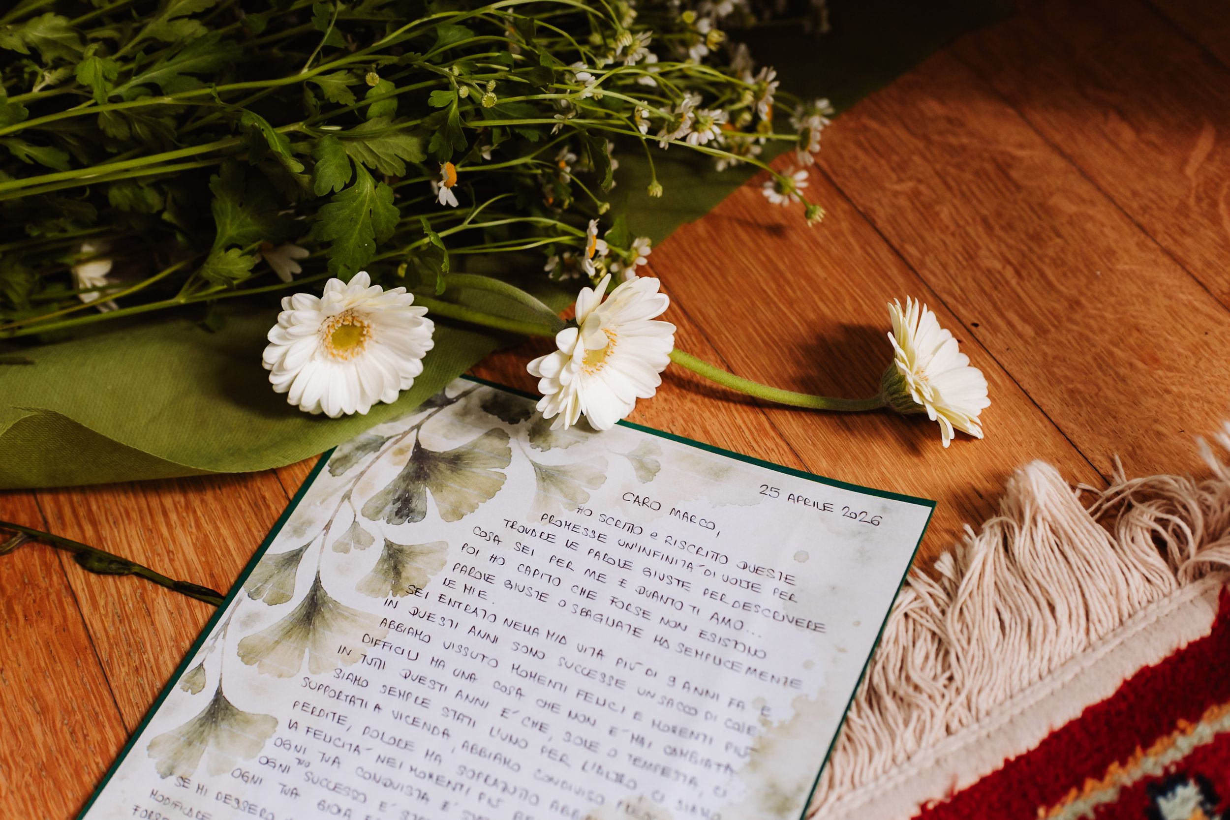 White daisies and wildflowers rest on a handwritten letter on a wooden floor beside a red fringed rug.