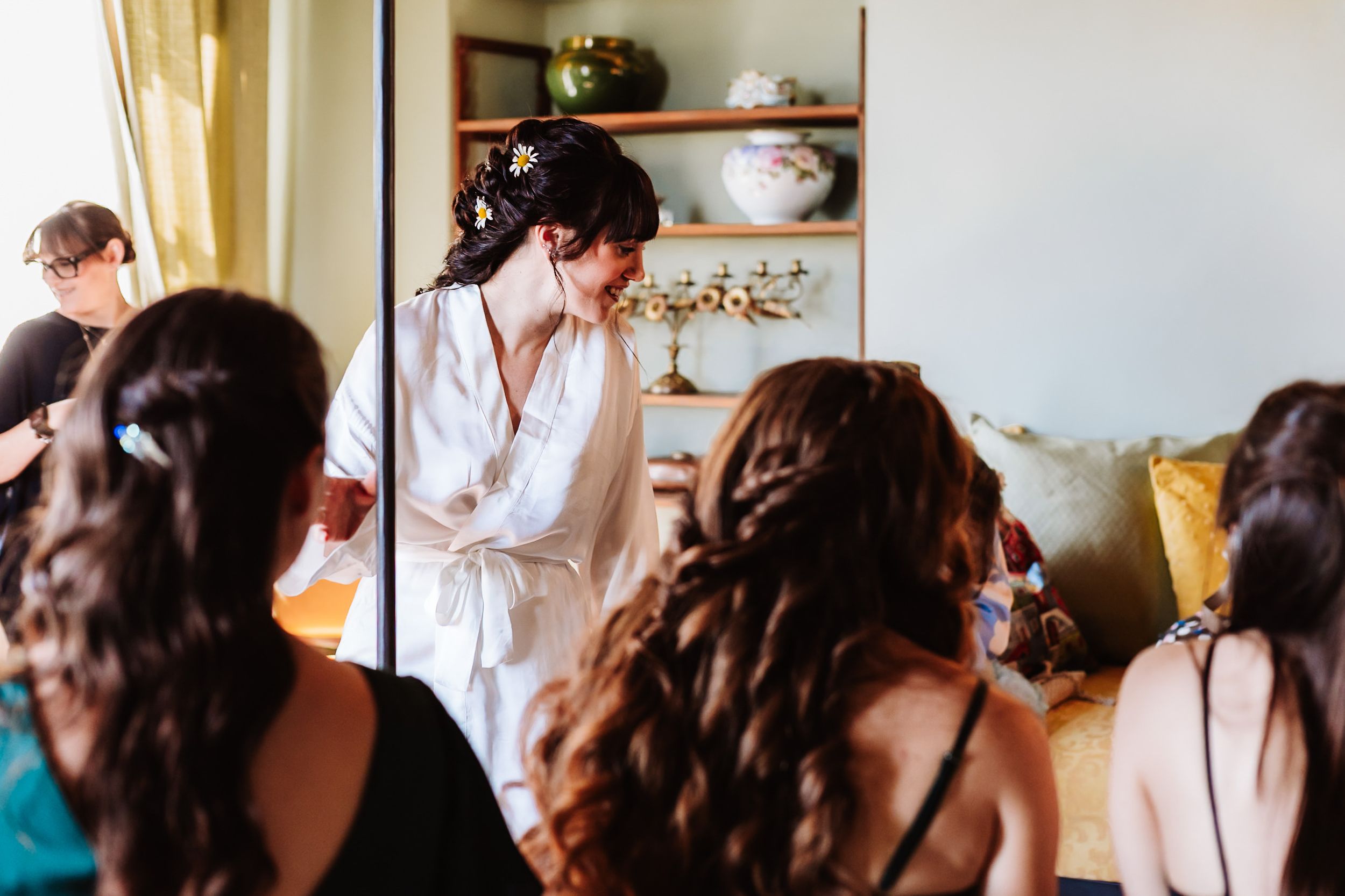 Bride in white robe getting ready with bridesmaids in a bright room before the wedding ceremony.