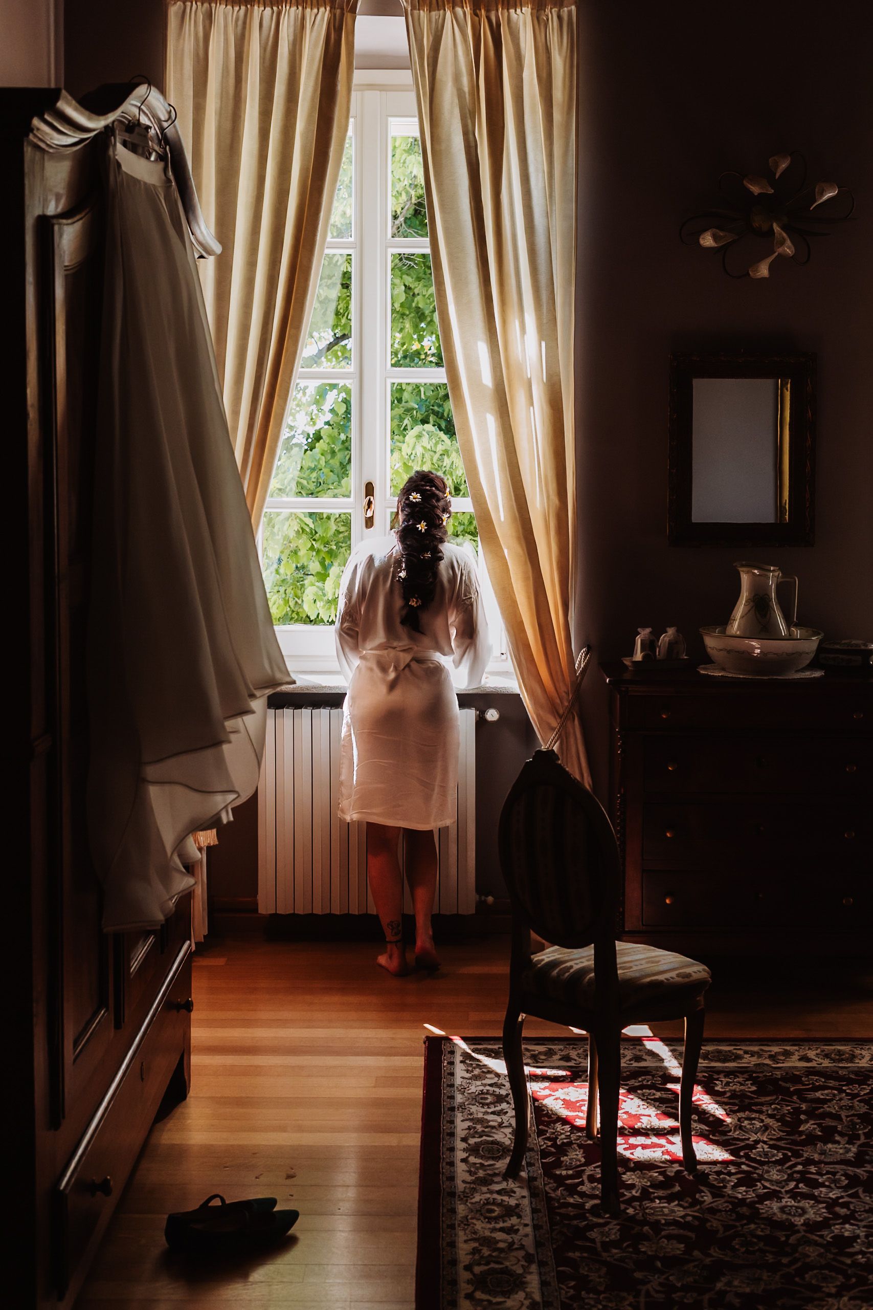 Bride in white robe gazing out a sunlit window, wedding dress hanging nearby in an elegant room.