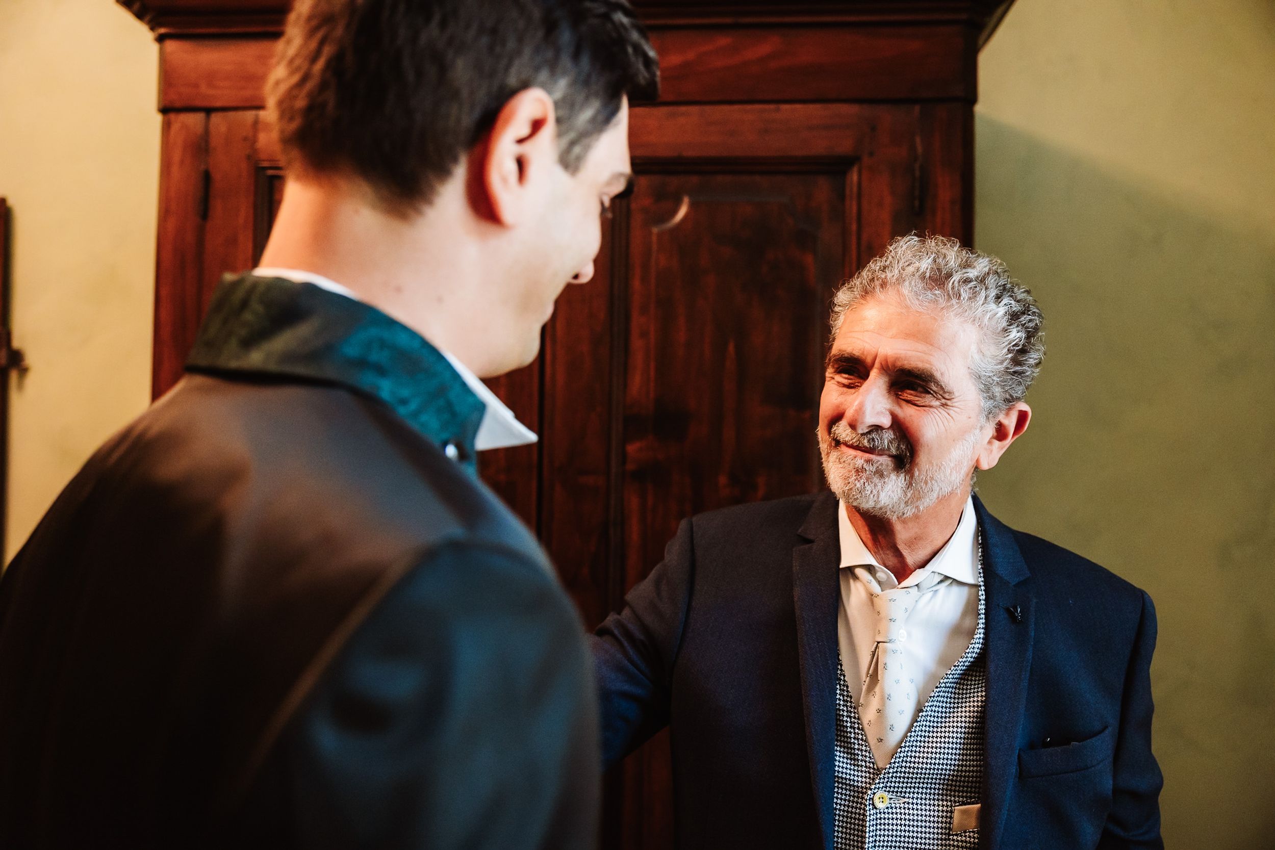 Two men in formal attire share a warm conversation near a wooden cabinet at a wedding event.